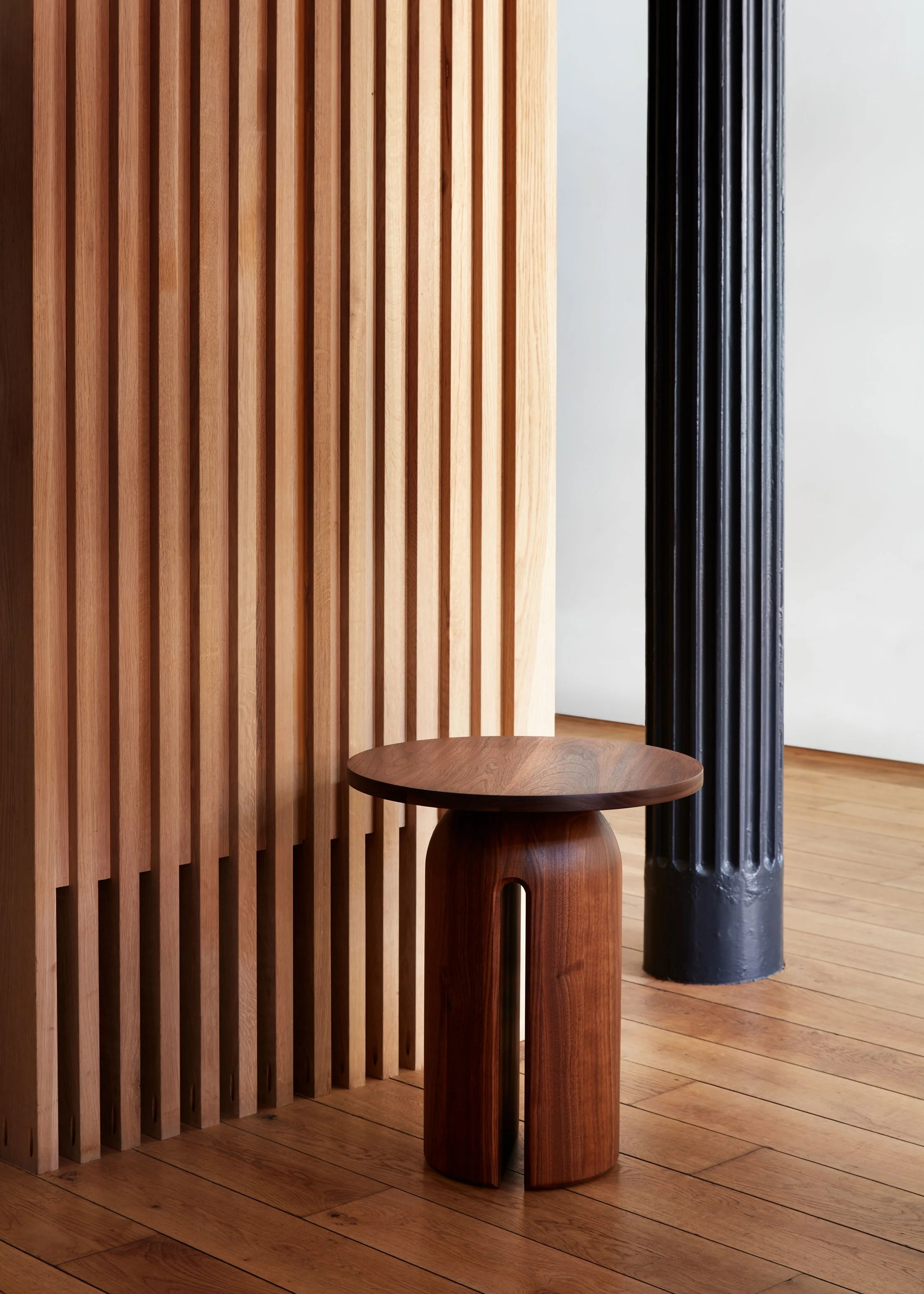 A minimalist wooden stool with a rounded top and wide, curved legs, placed on a polished wooden floor next to a textured wooden slat wall and a tall black fluted column.
