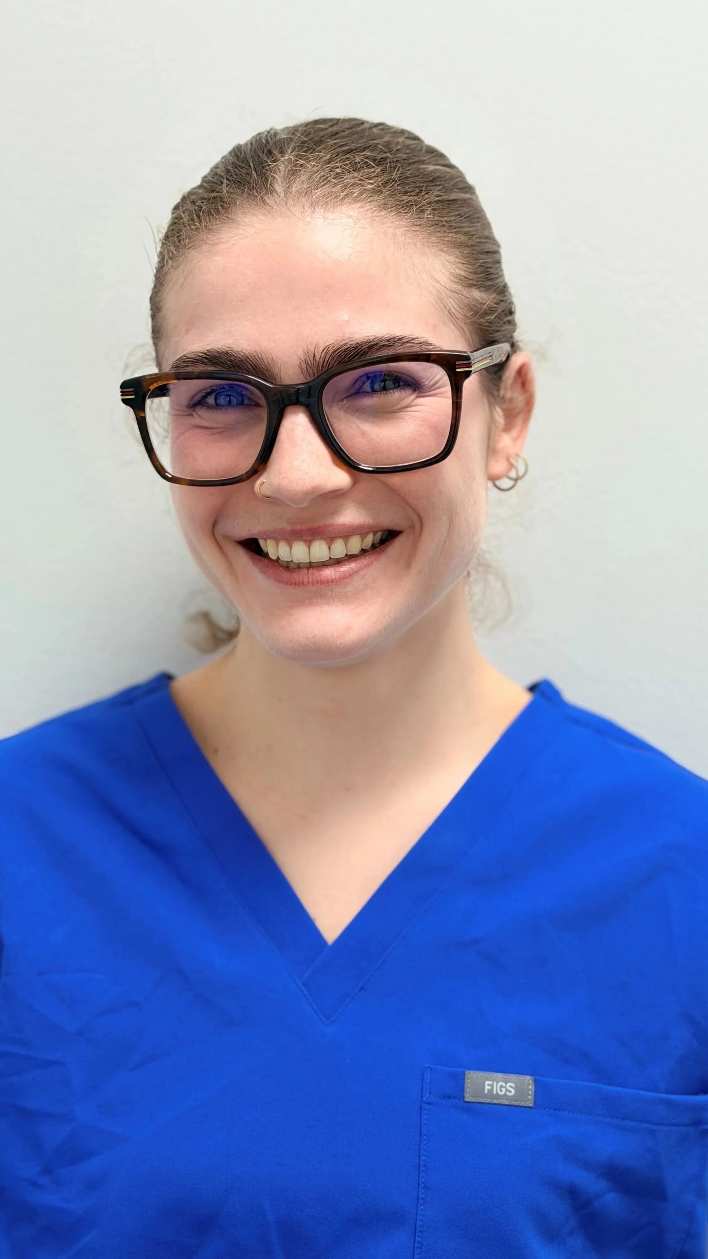 Een glimlachende vrouw in een blauwe medische uniform met een naamplaatje dat 'FIGS' zegt, draagt tortoise-omrande brillen en heeft een neusring, staand tegen een witte achtergrond.