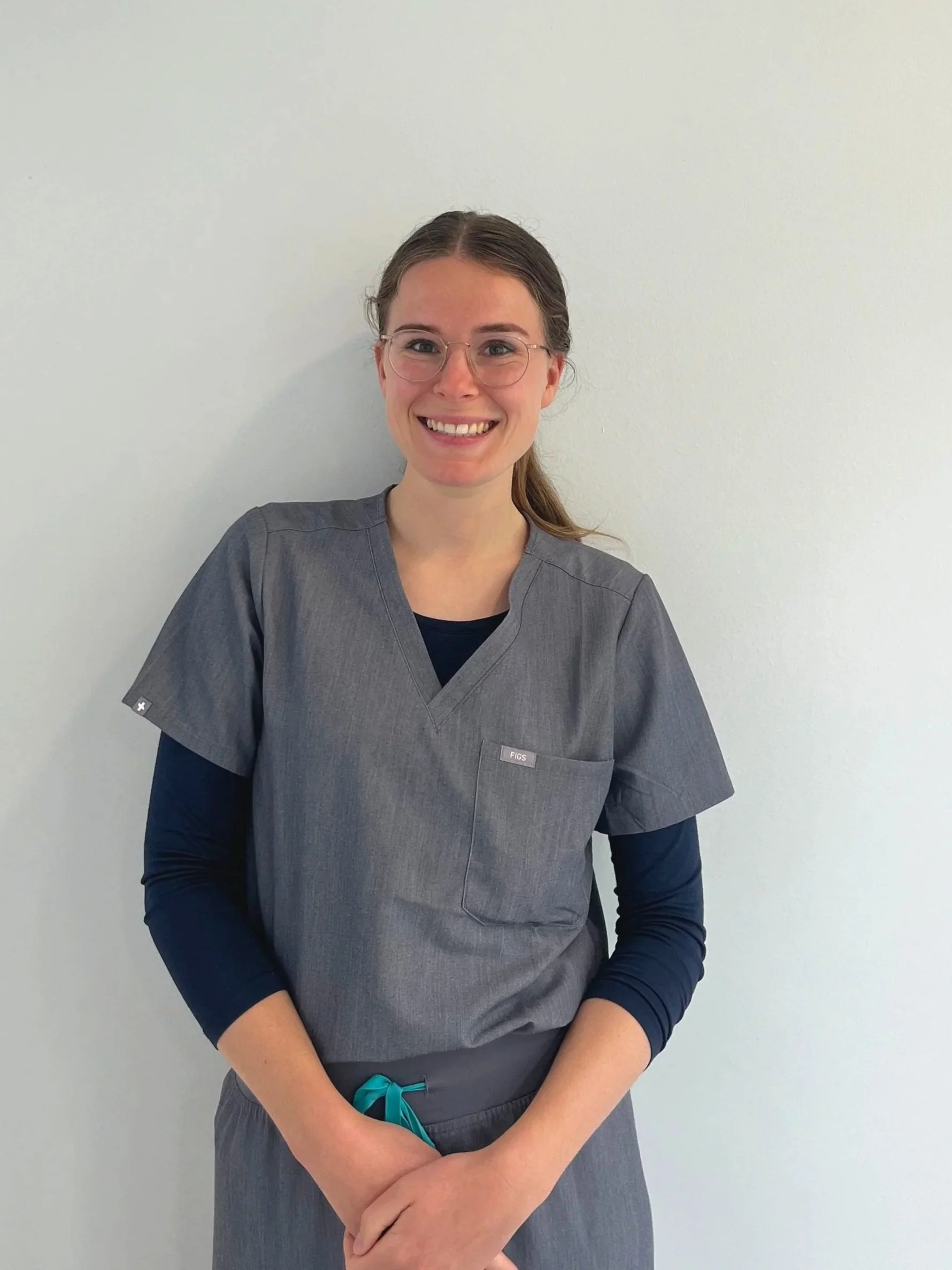 Person wearing gray scrubs and glasses, standing against a plain wall, smiling.