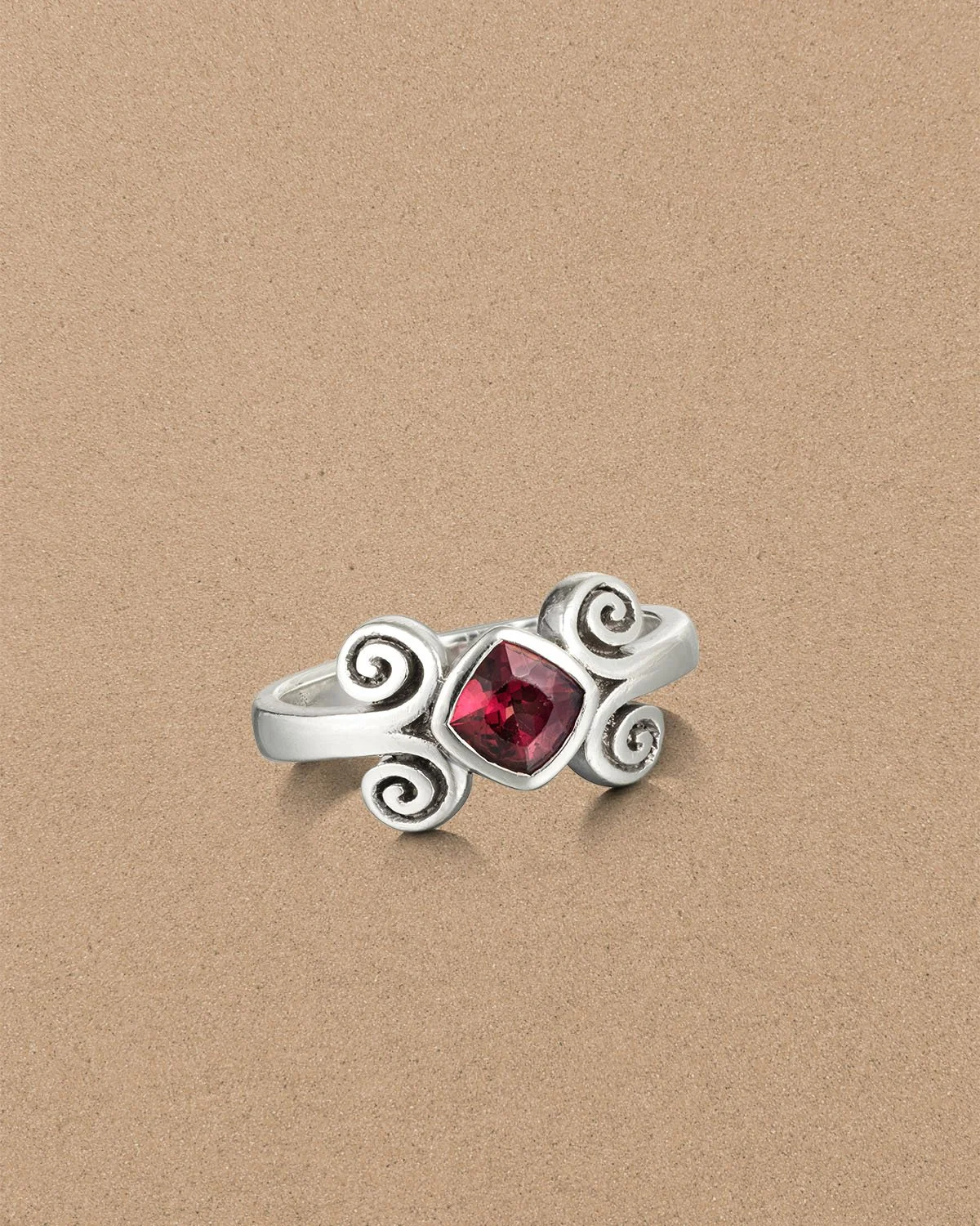 Silver ring with a square-cut red gemstone and decorative swirl designs, resting on a sandy surface.