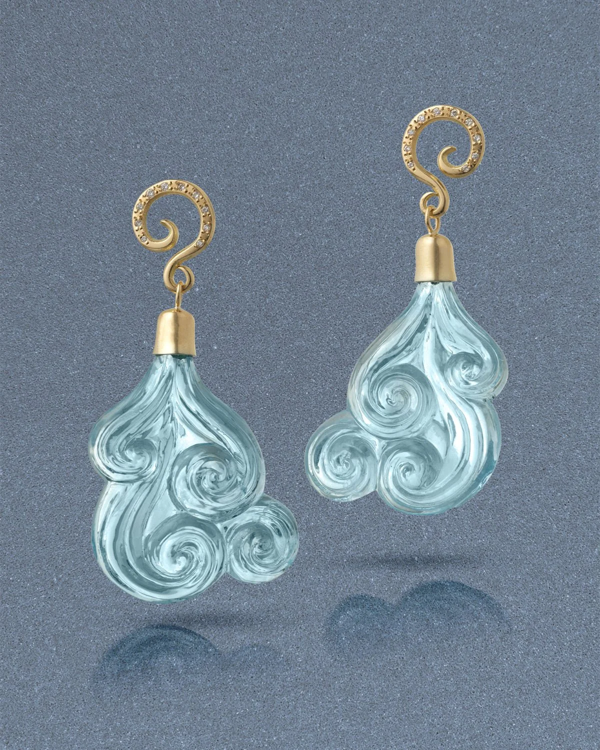 A pair of transparent glass earrings with swirling patterns, gold hooks, and small gemstones on the hooks, set against a gray background.