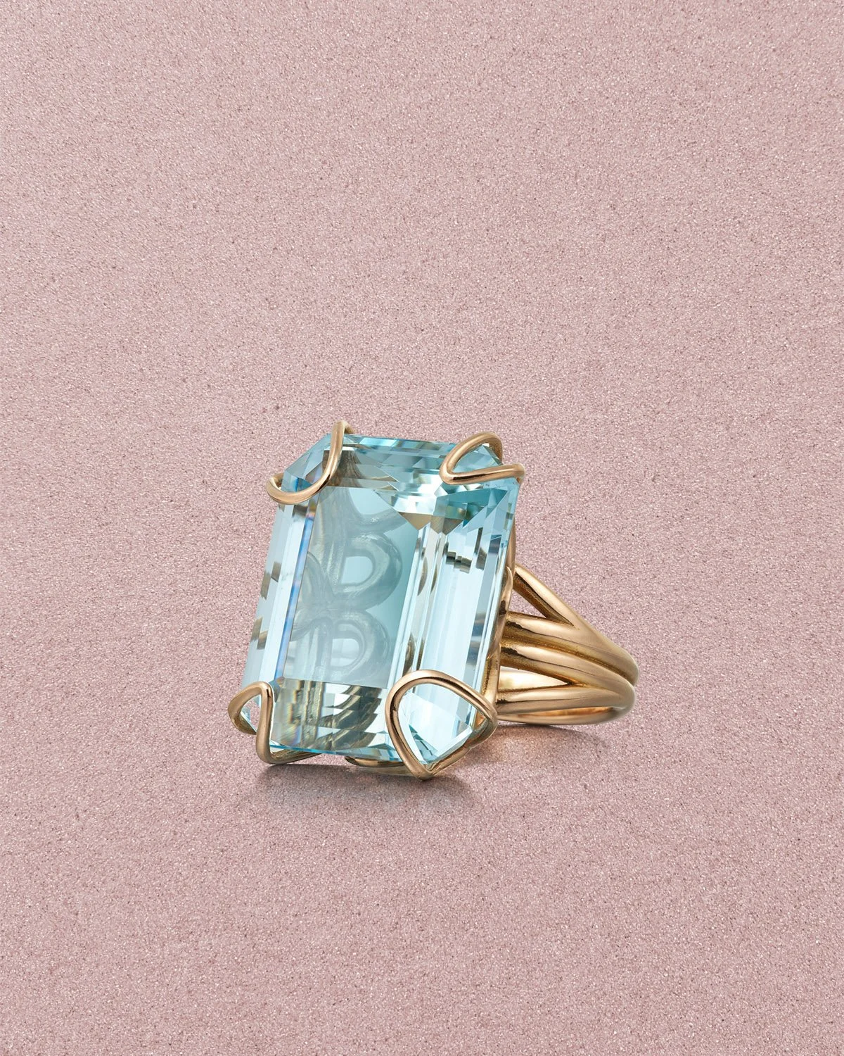 A gold ring with a large rectangular light blue gemstone set in a unique prong setting on a pink surface.