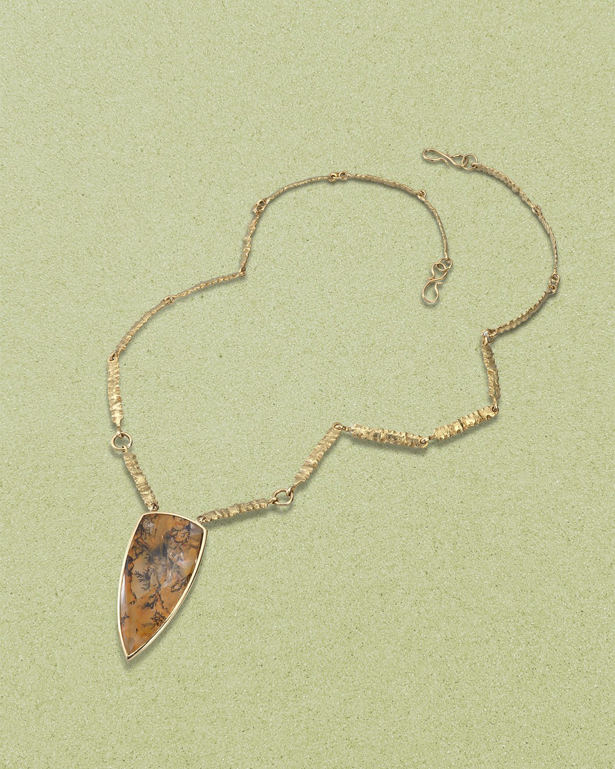 Gold necklace with a large, leaf-shaped pendant containing a patterned brown and black stone, lying on a light green surface.