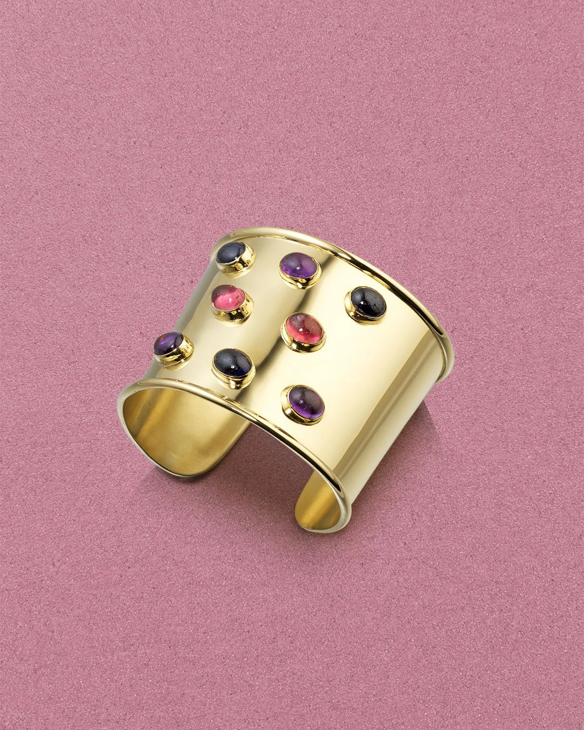 A gold wide cuff bracelet decorated with small pink, purple, and black stones, placed atop a pink textured surface.