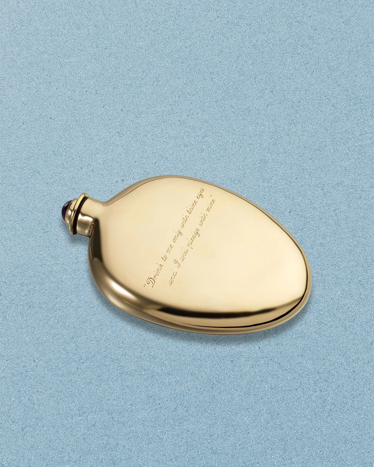 A gold whiskey flask, shaped like a teardrop placed on a blue textured background, with a personalised inscription on its surface.