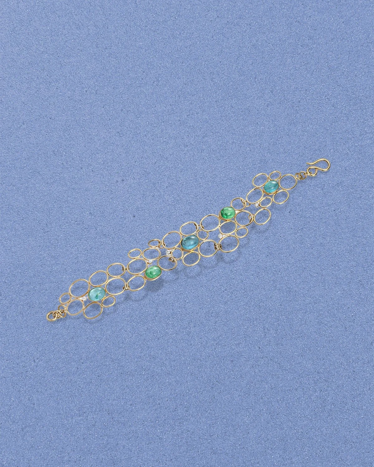 Gold bracelet, inspired by ocean bubbles with circular links and blue and green stones on a blue textured surface.