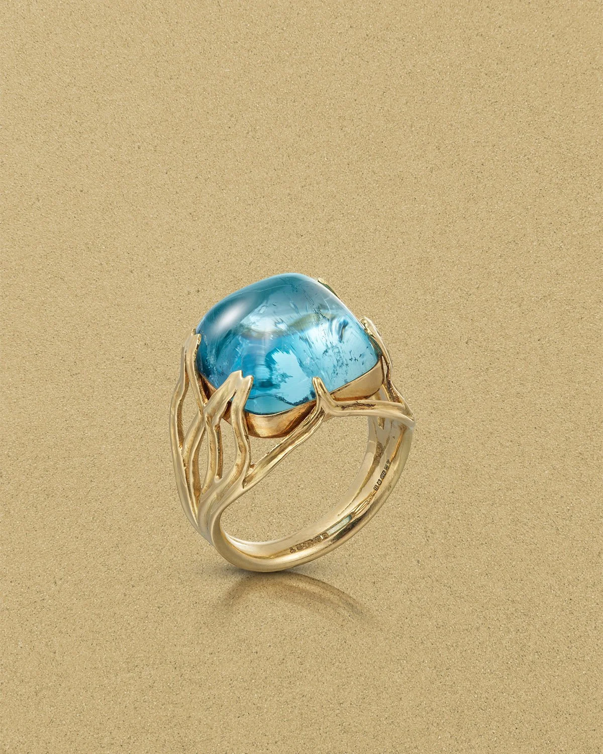 Gold ring with a large, blue oval gemstone set in an intricate, tree branch-like design against a beige background.