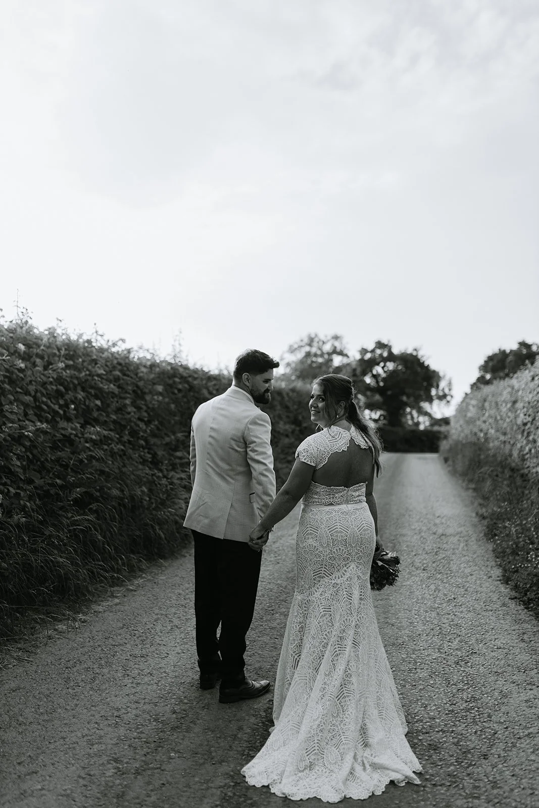 A bride and groom holding hands and walking down a rural gravel path, looking at each other, with bushes on either side and a cloudy sky above.