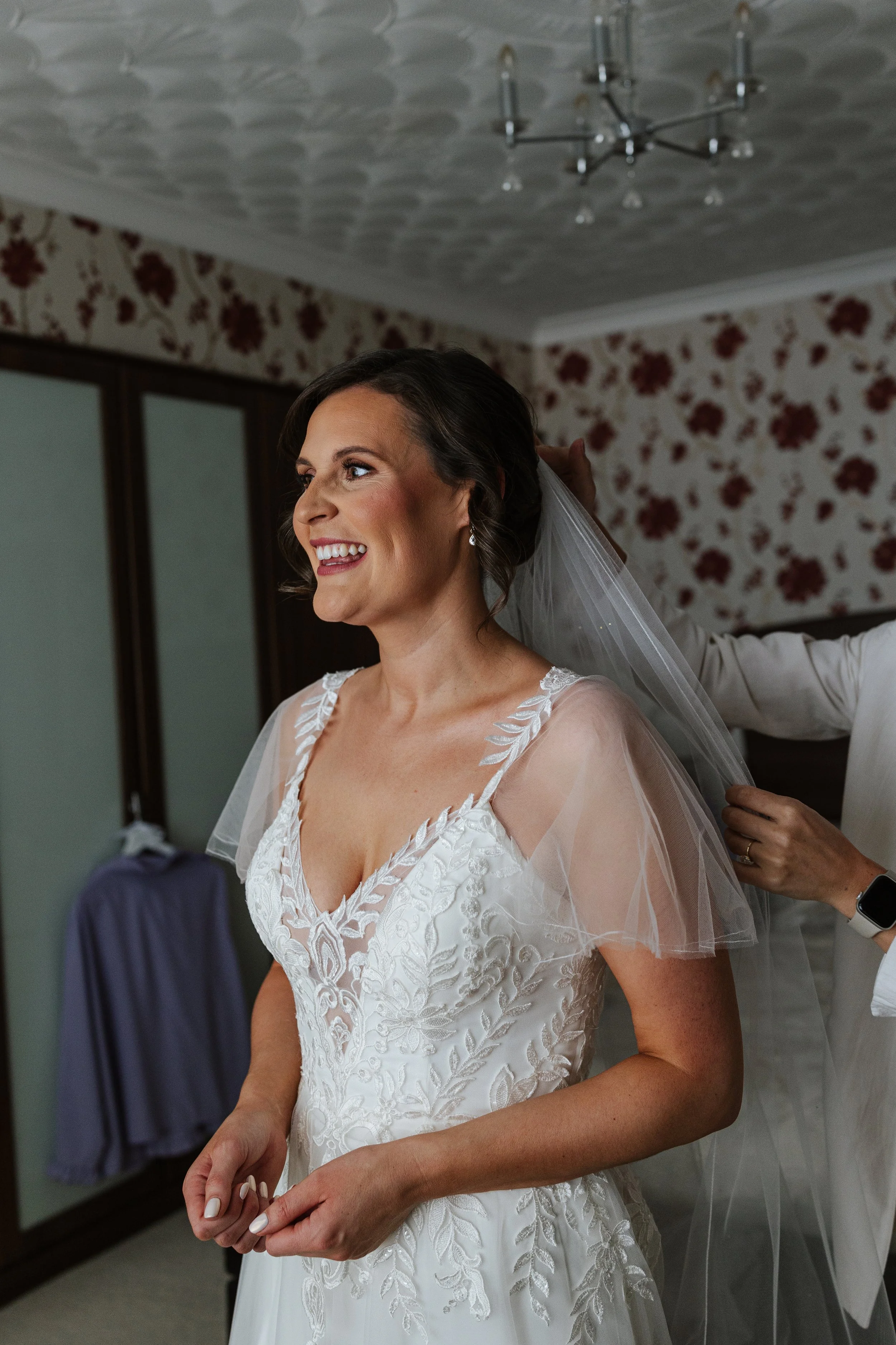 A bride is smiling and wearing a white wedding dress with lace details, as someone adjusts her veil in a room with floral wallpaper.