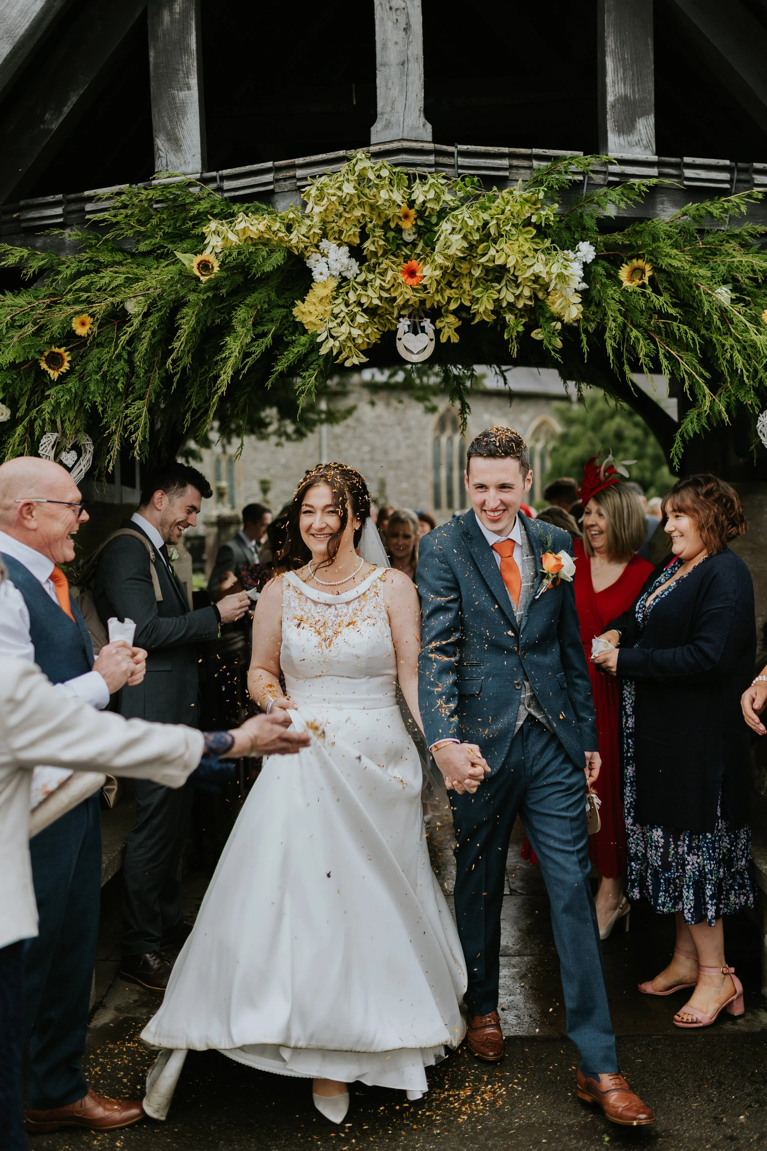 A bride and groom holding hands and smiling as they walk under a floral arch at their wedding. Friends and family surround them, celebrating outside near a church.