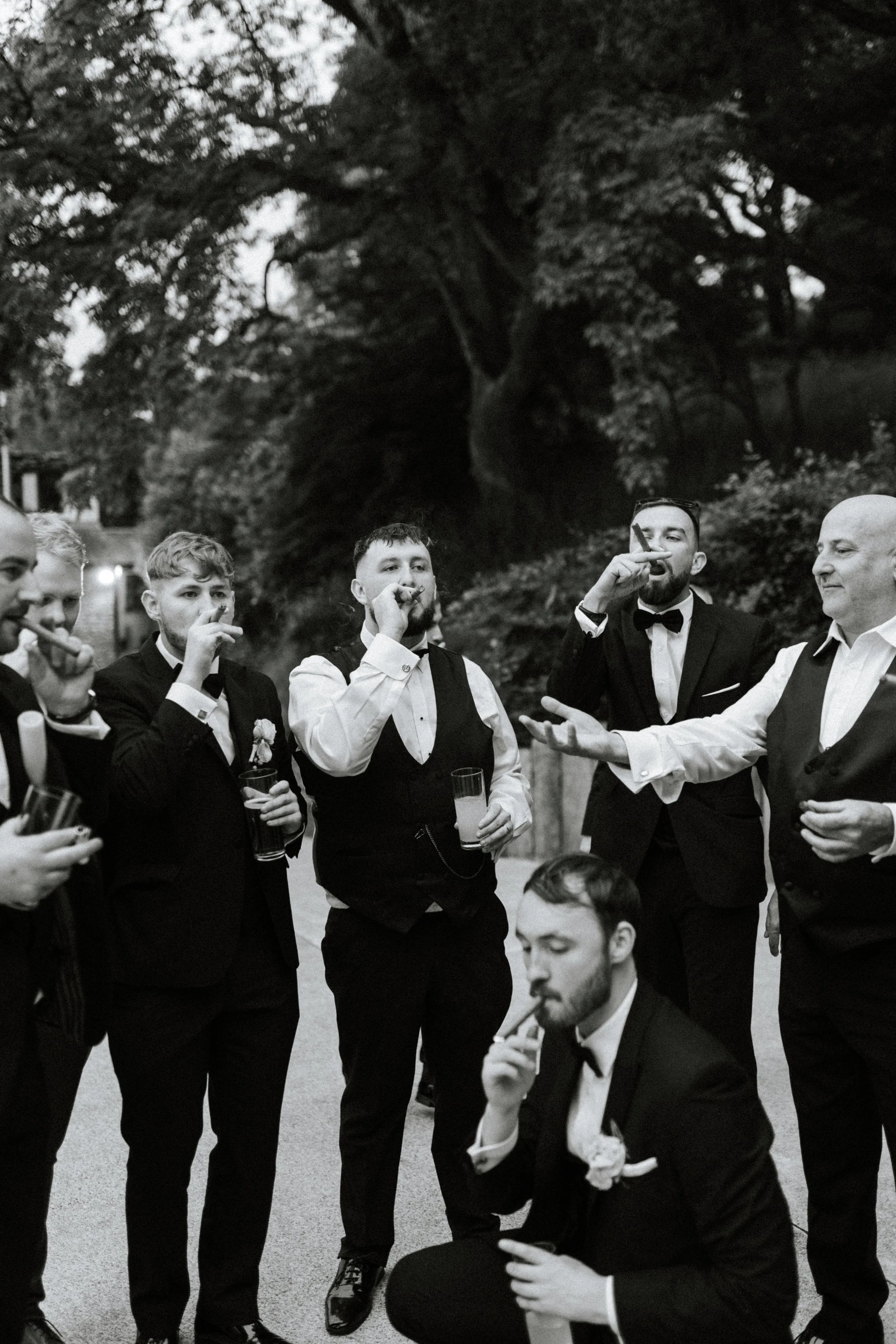 A group of men dressed in tuxedos and bow ties gathering outside under trees, smoking cigars and drinking beverages.
