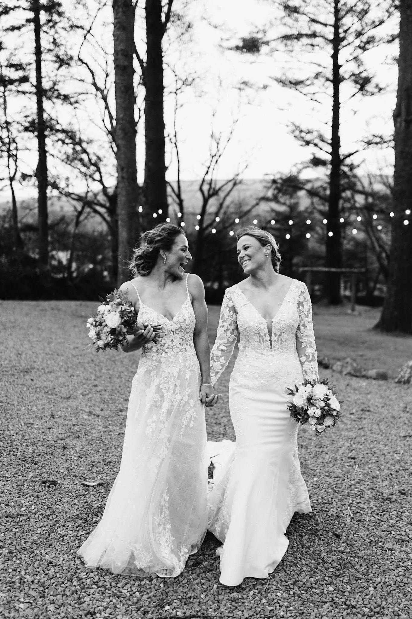 Two women in wedding dresses holding hands and smiling at each other outdoors, with trees and string lights in the background.