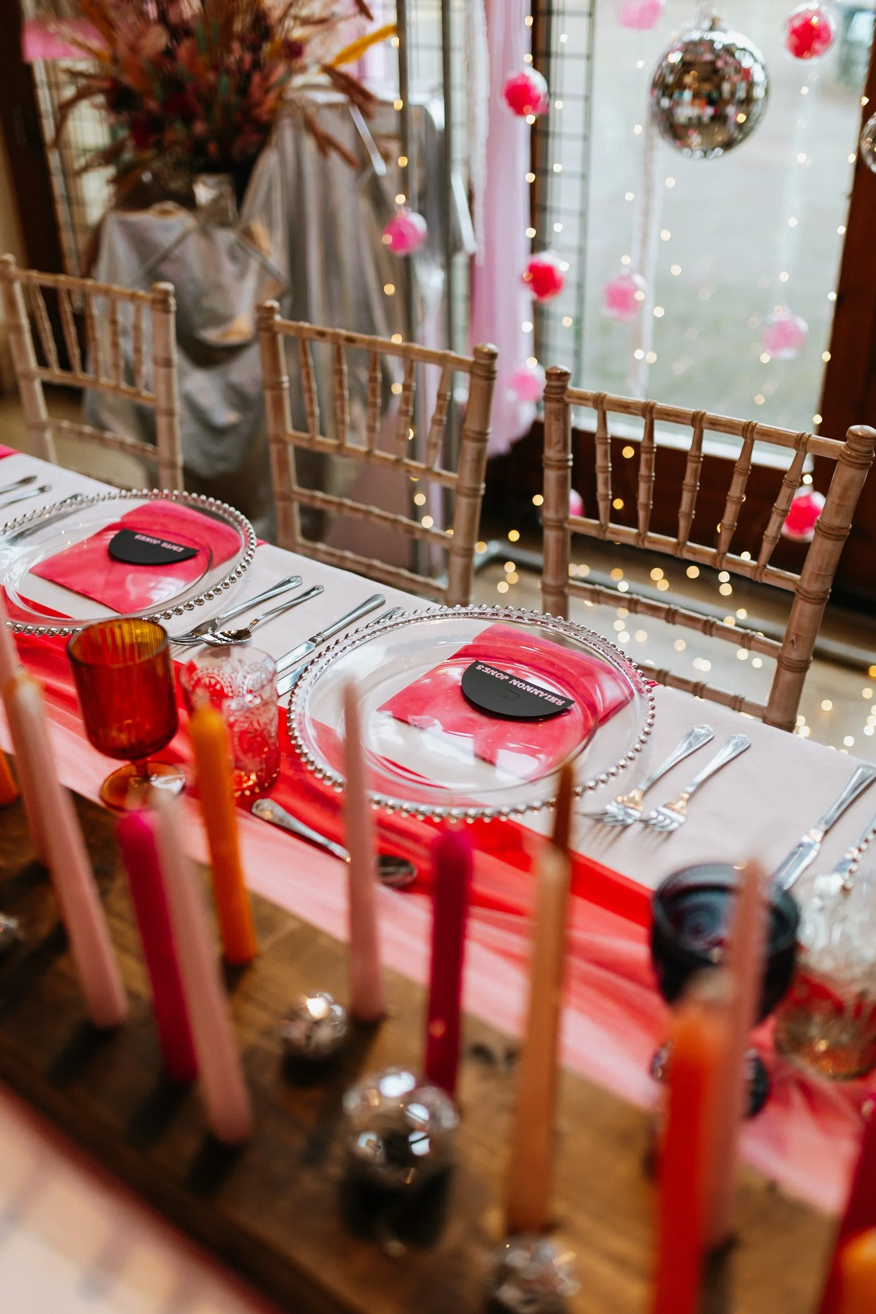 Table setting for a celebration with pink and red decorations, including plates, glasses, and napkins, with a window and hanging decorations in the background.