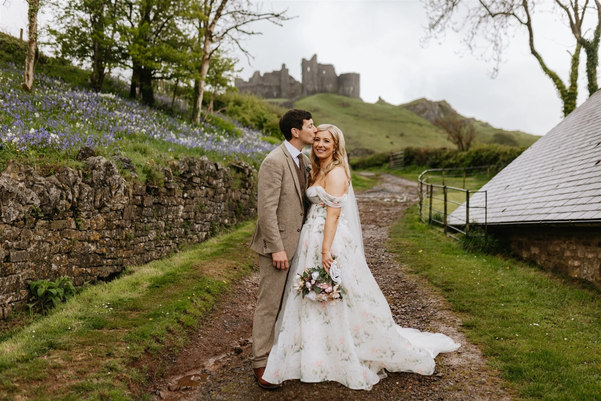 A couple dressed in wedding attire stand on a dirt path in a rural setting, with a stone wall on one side and a green grassy hill with flowers on the other. The groom is kissing the bride's forehead, and she is smiling while holding a bouquet. In the