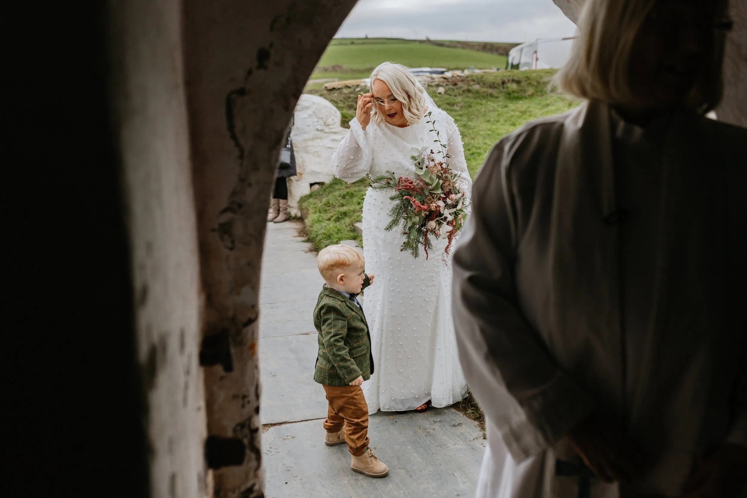 A woman with blonde hair in a white dress holding a bouquet of flowers, standing outdoors near a child and adult, as seen through a circular opening in a rustic wooden structure, with green rolling hills in the background.