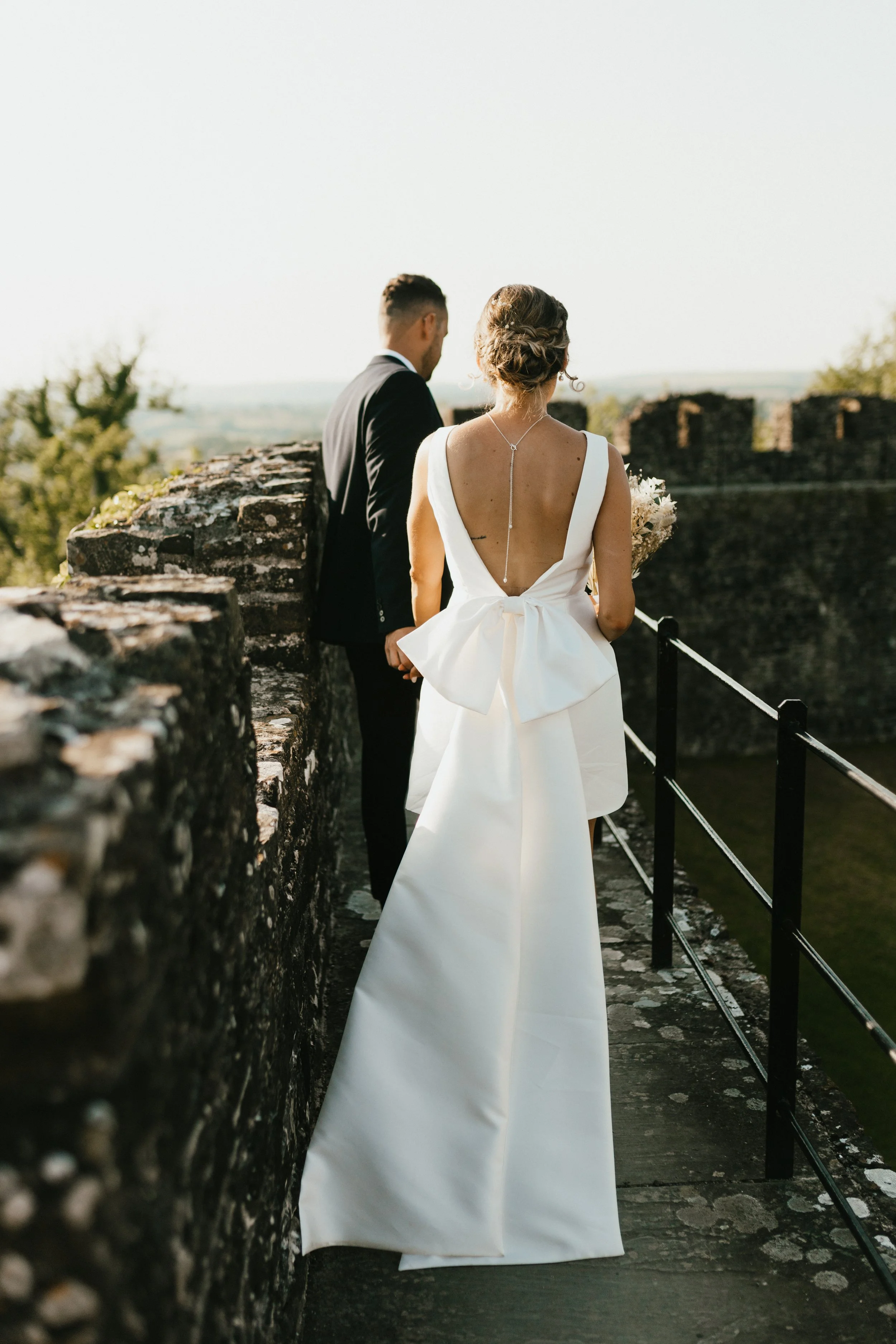 A bride and groom holding hands and standing on a stone pathway or terrace with ancient stone walls, overlooking a scenic landscape, during a wedding celebration.