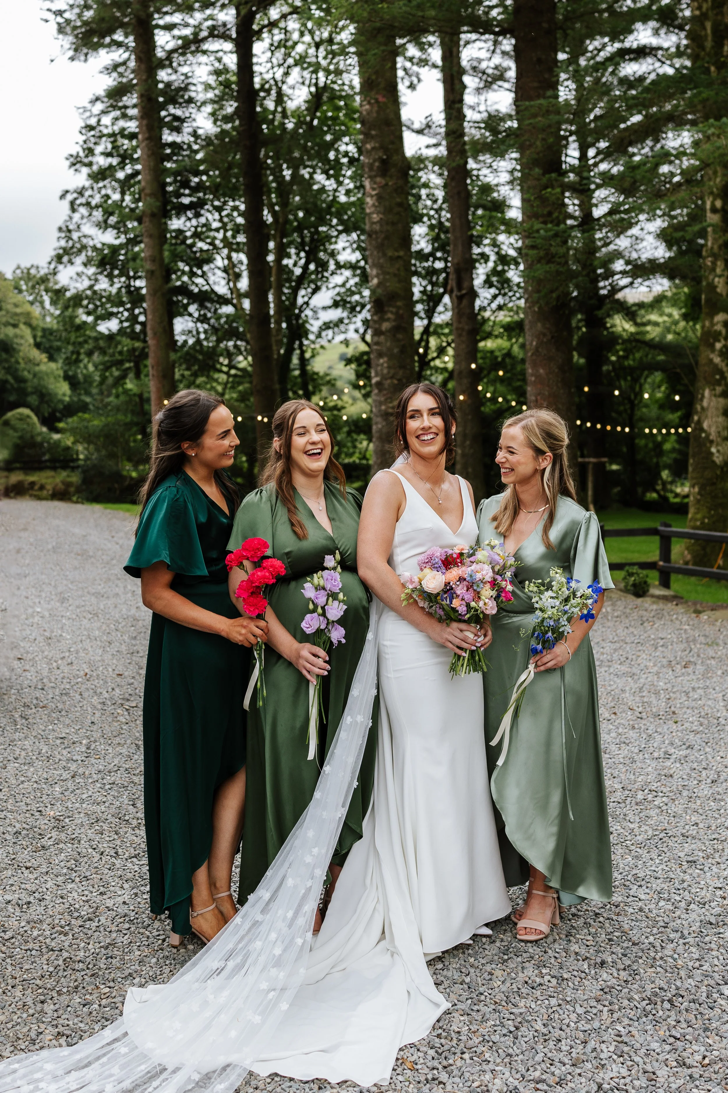 Four women at a wedding outdoors in a wooded area. The bride, in a white gown, holds a bouquet and stands in the middle. Three bridesmaids in green dresses hold smaller bouquets and stand on either side. They are smiling and looking at each other. St