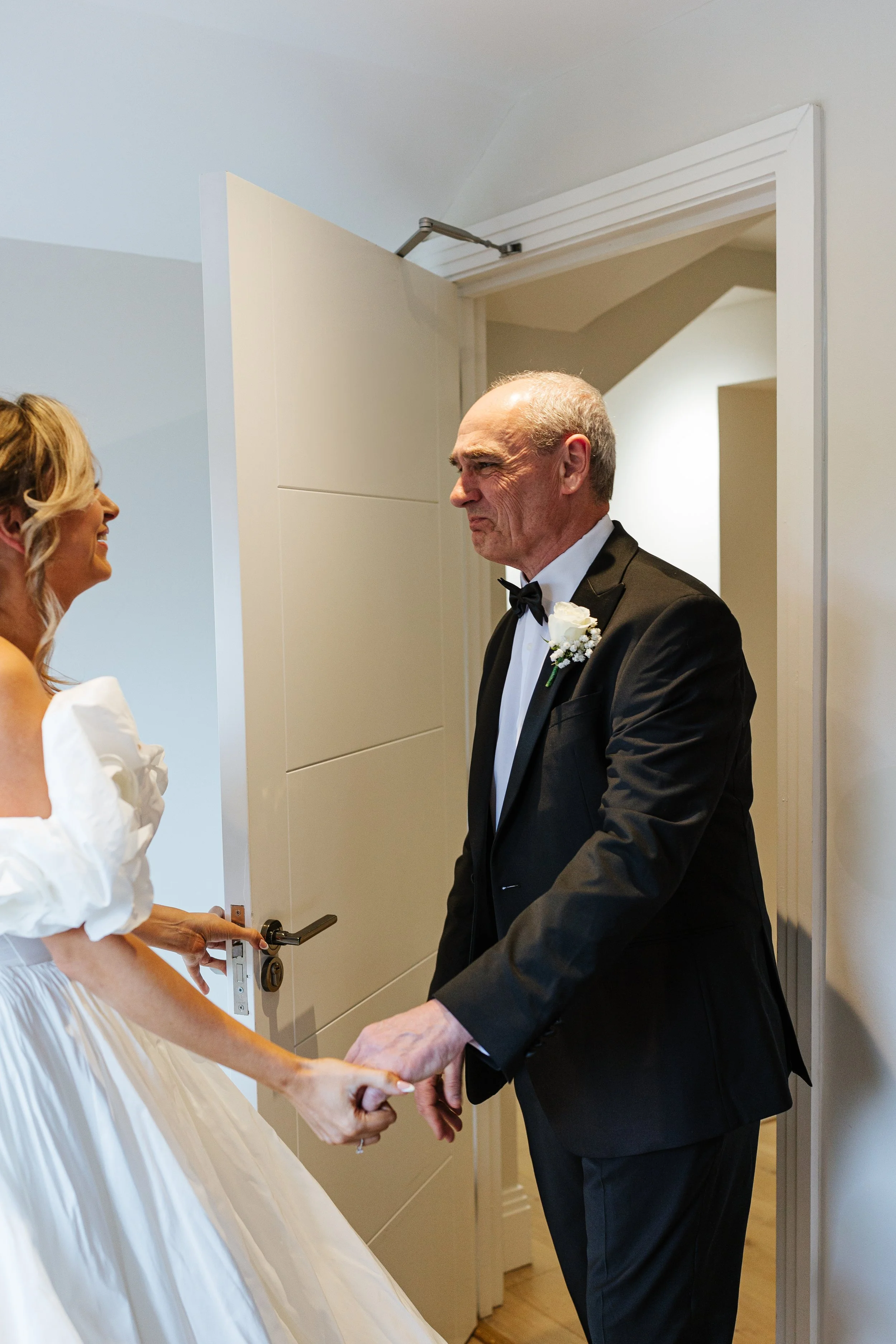 A bride and an older man, possibly her father, are holding hands in front of an open door. The bride is smiling and wearing a white dress with puffy sleeves, and the man is wearing a black tuxedo with a bow tie and a white boutonniere. They are about