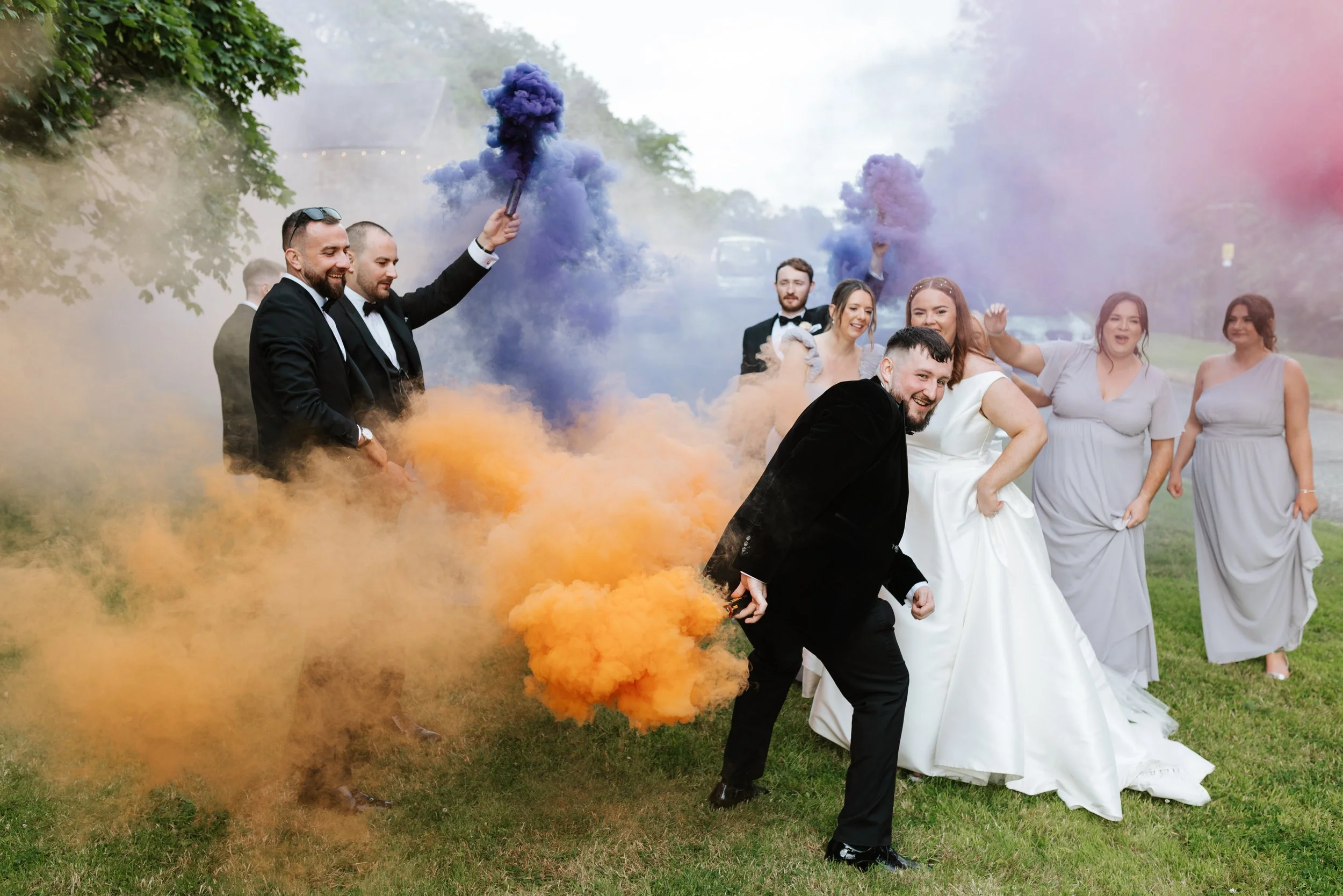 A group of wedding guests in formal attire celebrating outdoors, with many holding purple and orange smoke bombs, dispersing colorful smoke in the air.