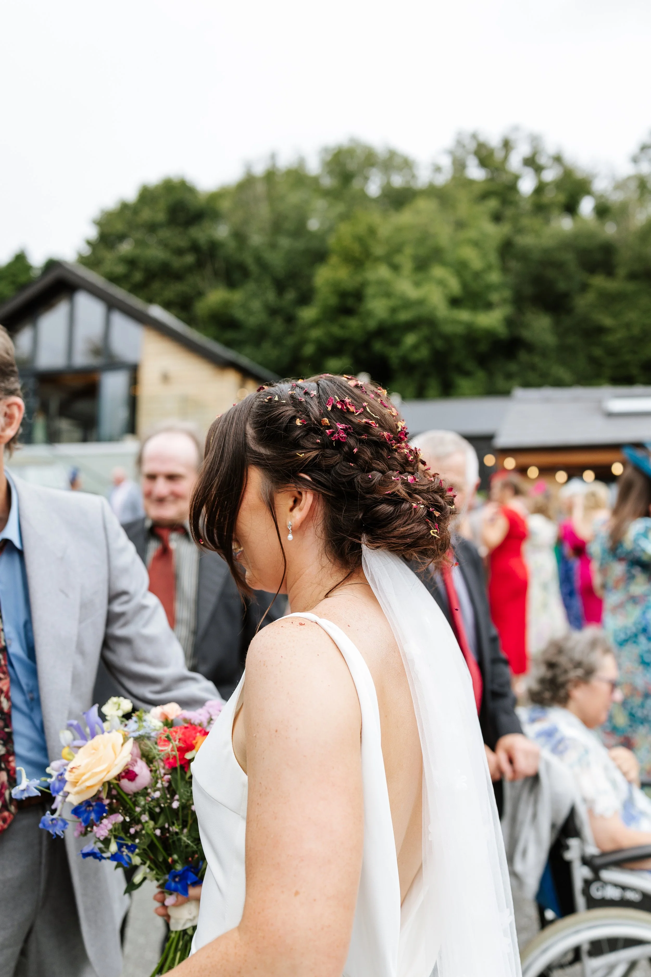 A woman with brown hair in an updo, decorated with pink flower petals, wearing a white dress, is at a gathering outdoors. She is holding a bouquet of flowers while people in formal attire are present in the background, with some trees and buildings v