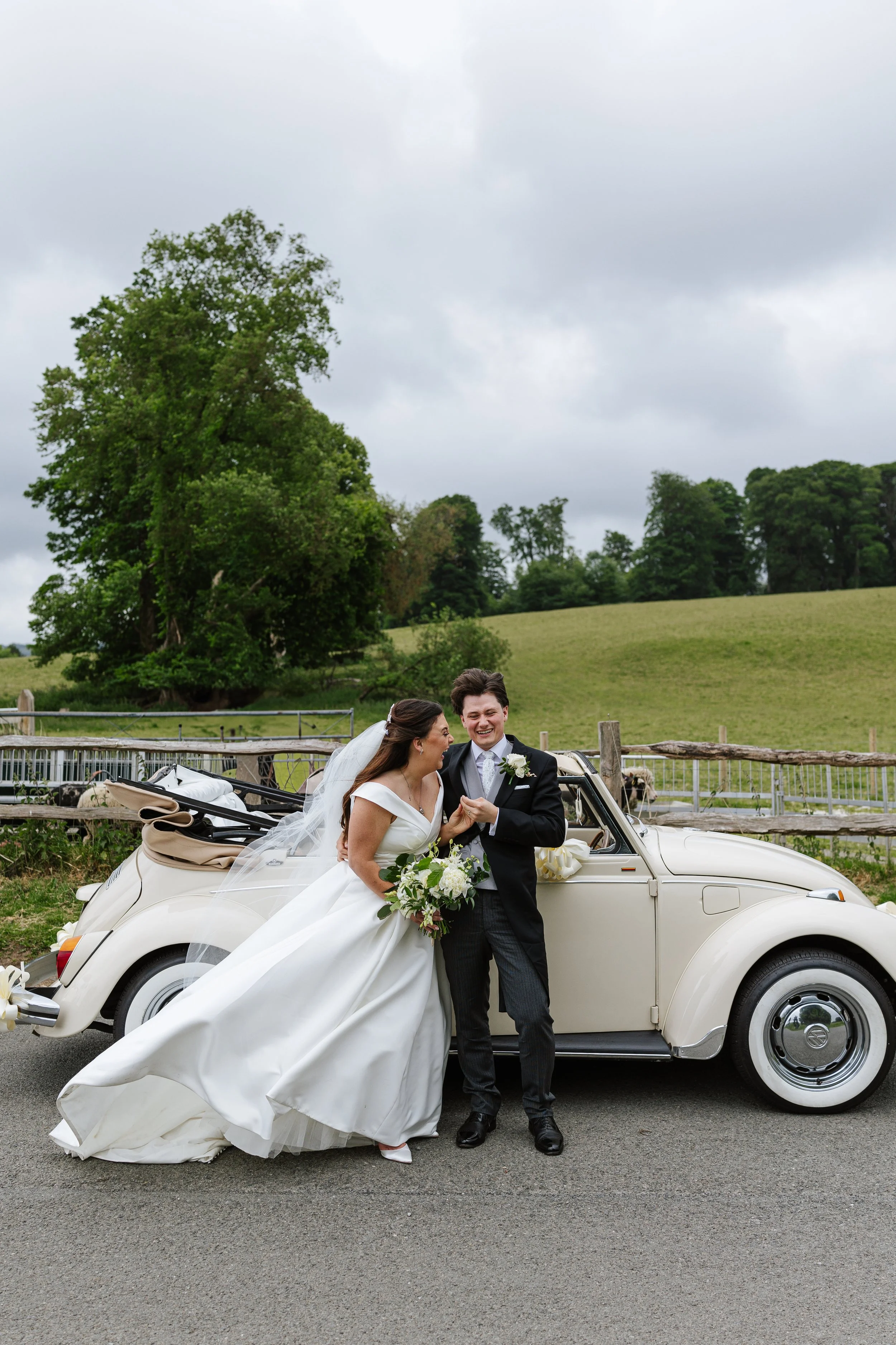 A bride and groom standing beside a vintage white convertible car with a mountainous and cloudy sky background. The bride is in a white wedding gown holding a bouquet, and the groom is in a black suit with a boutonnière.