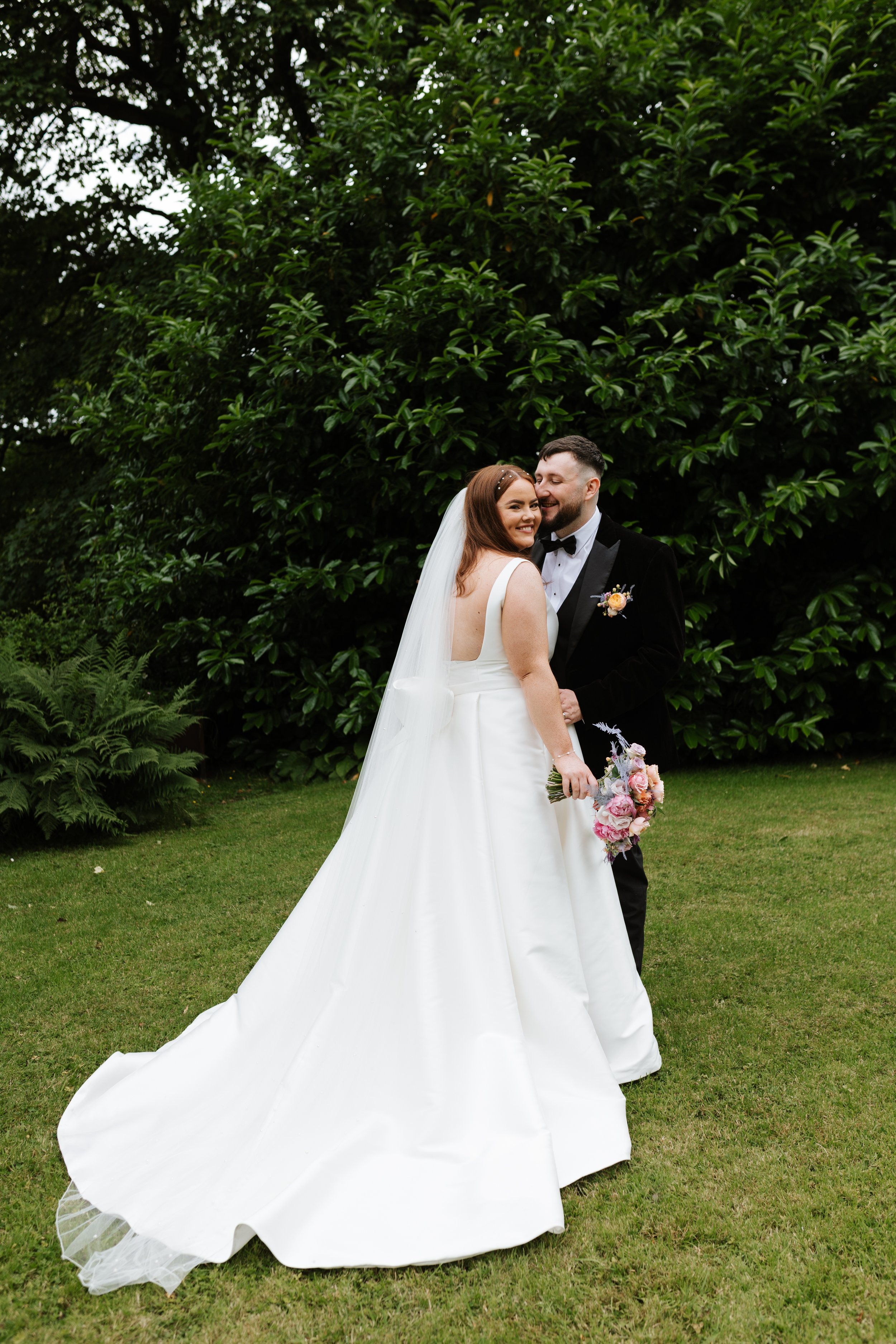 A bride and groom standing on grass in front of large green trees, happy and smiling, with the bride holding a bouquet of pink and white flowers.