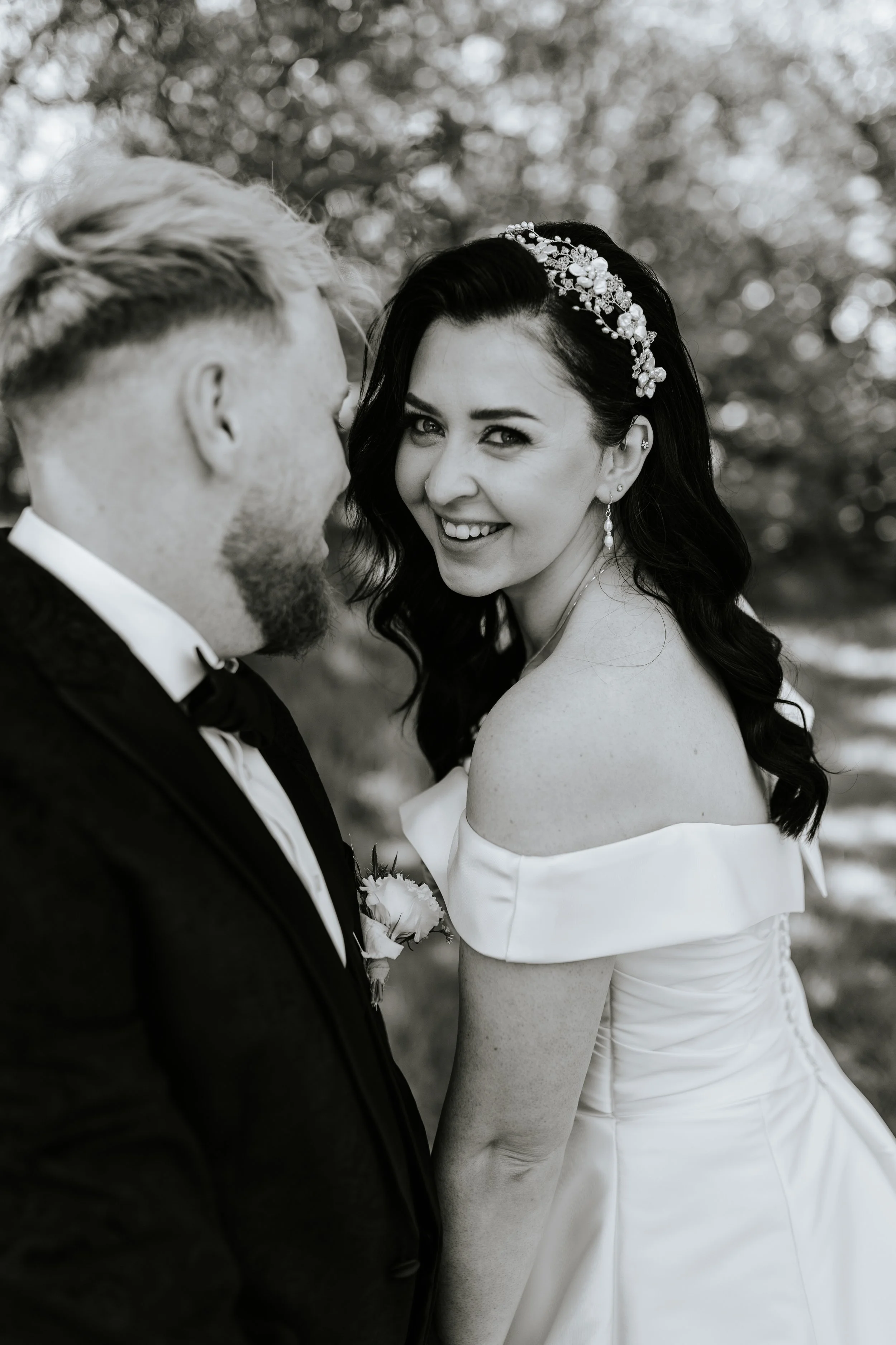 Black and white photo of a bride and groom outdoors, close-up view. The bride has dark hair with a floral headpiece and earrings, and the groom has light hair with a beard, wearing a tuxedo. They are smiling and gazing at each other.