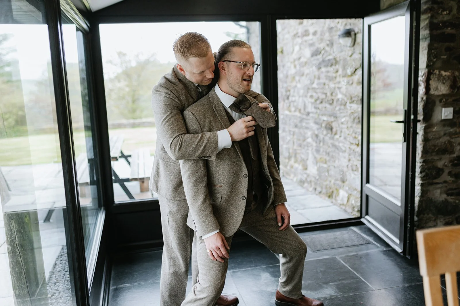 Two men wearing suits, one with glasses, engaging in a playful encounter inside a modern house with glass doors and stone walls, with outdoor greenery visible.