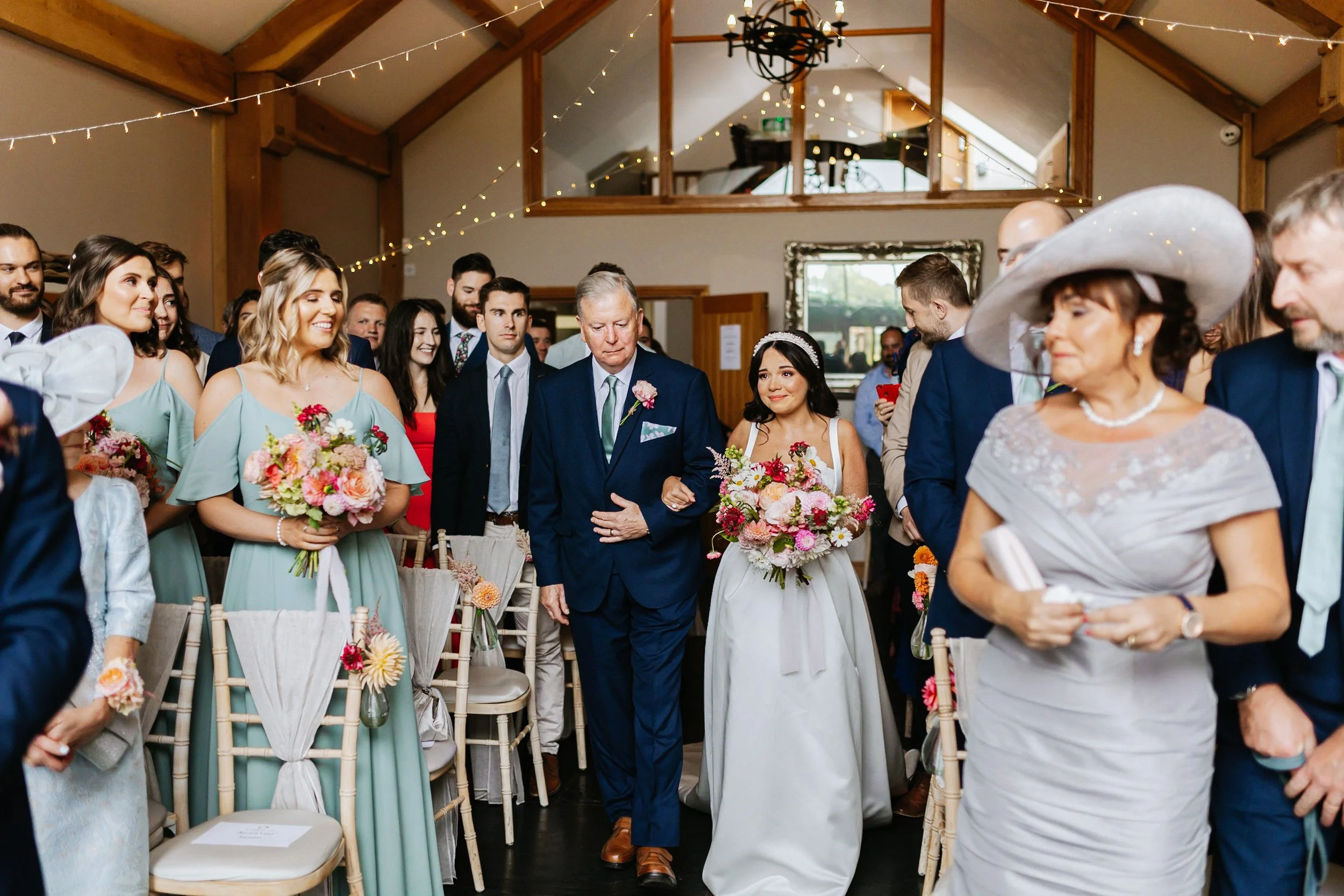 A diverse group of wedding guests, including women in pastel dresses and men in suits, are attending a wedding ceremony inside a rustic venue decorated with string lights. The bride, in a white dress and floral crown, is walking down the aisle with a