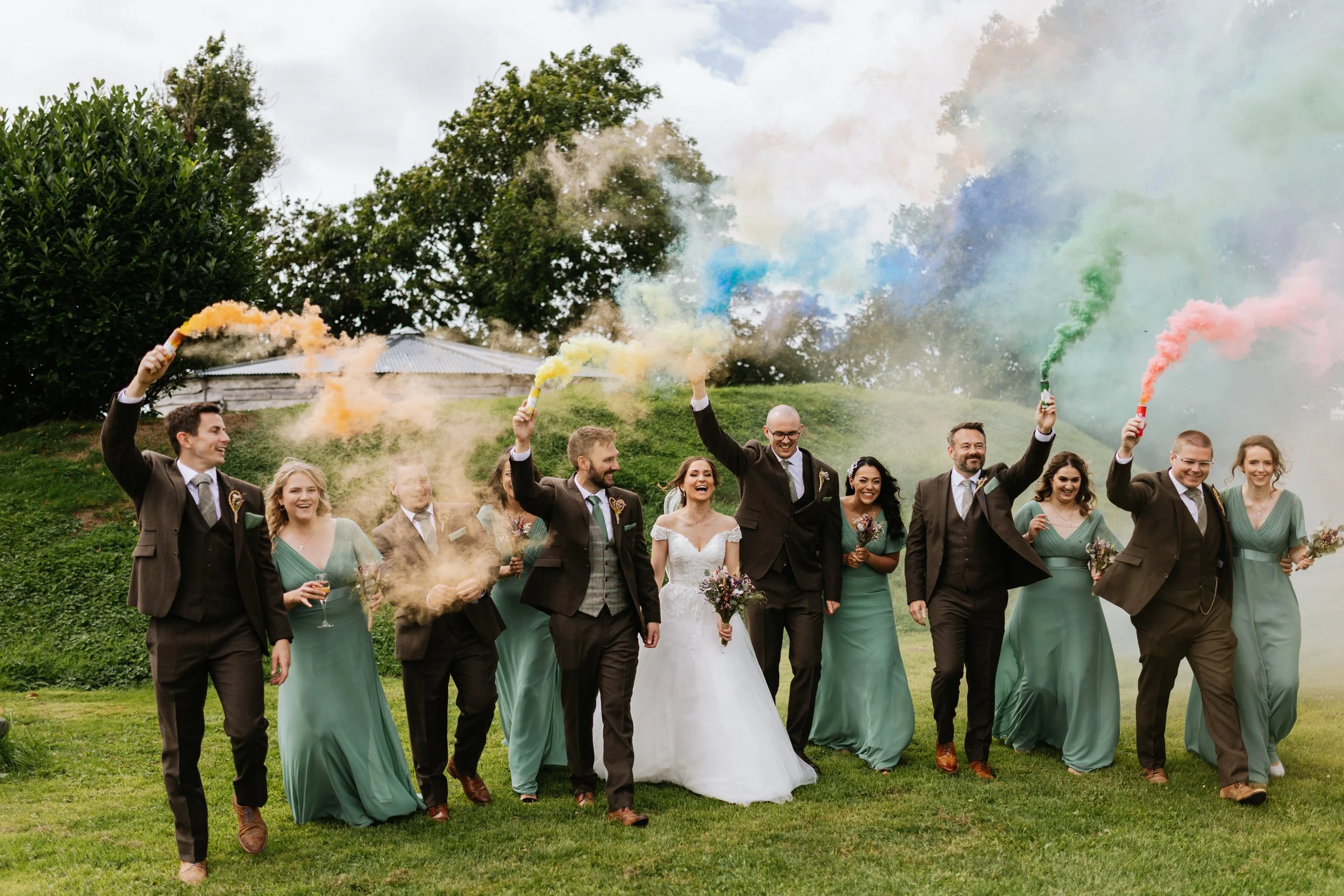 A wedding party walking outdoors on grass, holding colorful smoke flares.