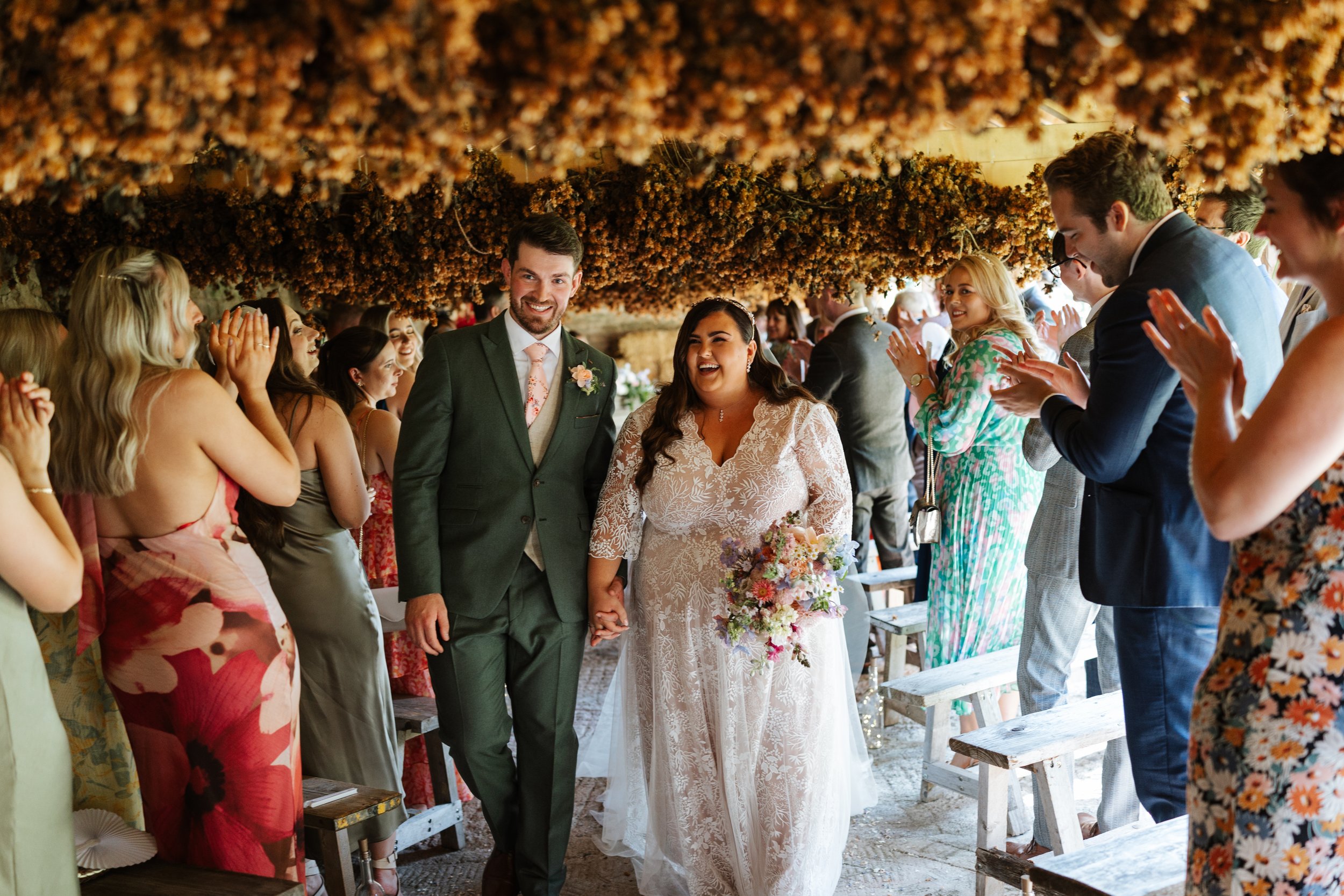 A wedding ceremony with a bride and groom walking hand in hand down the aisle, surrounded by seated and standing guests in an indoor setting decorated with hanging dried flowers.