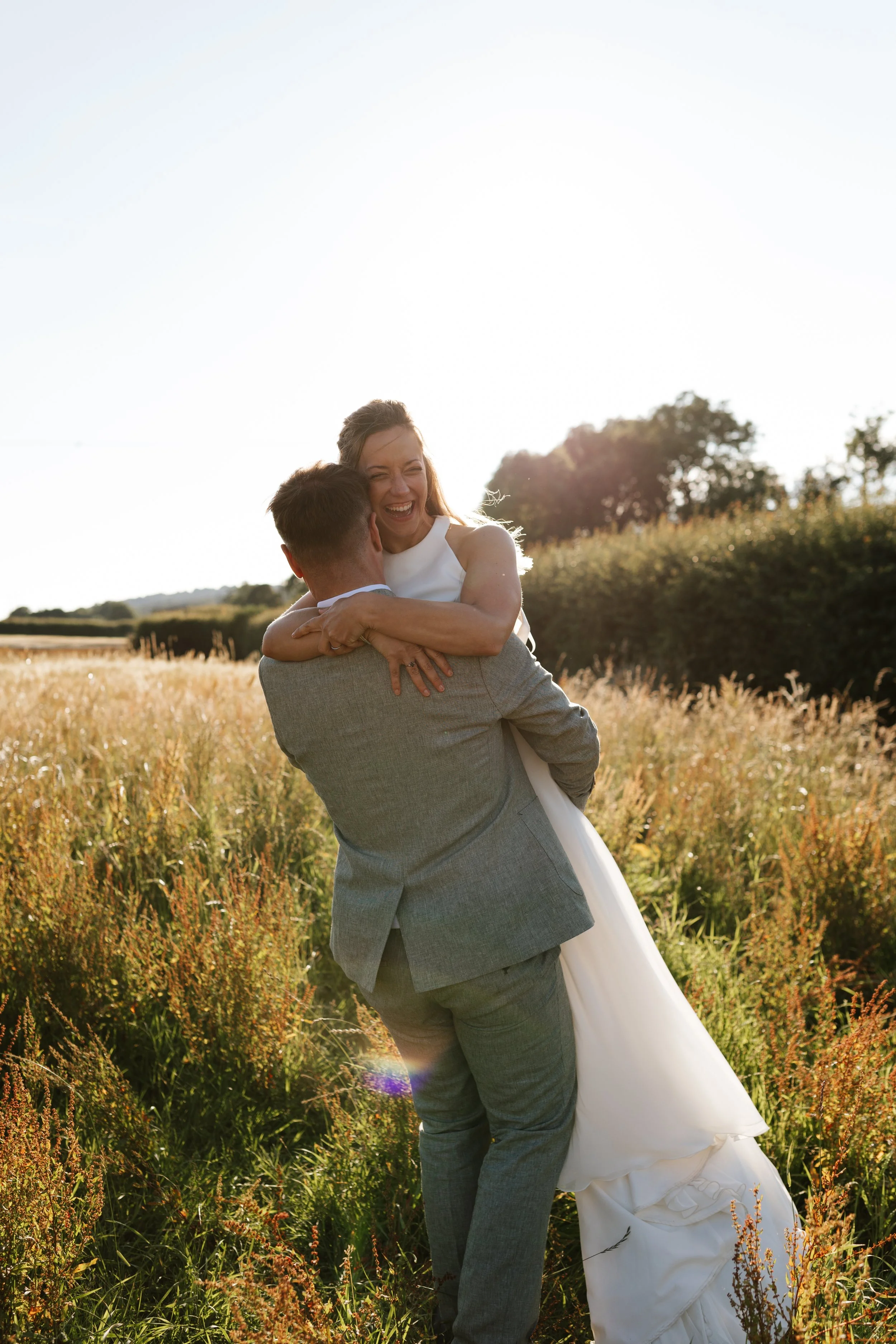 A couple, the man in a gray suit and the woman in a white dress, embracing and smiling happily in a sunlit field of tall grass and wildflowers during sunset.