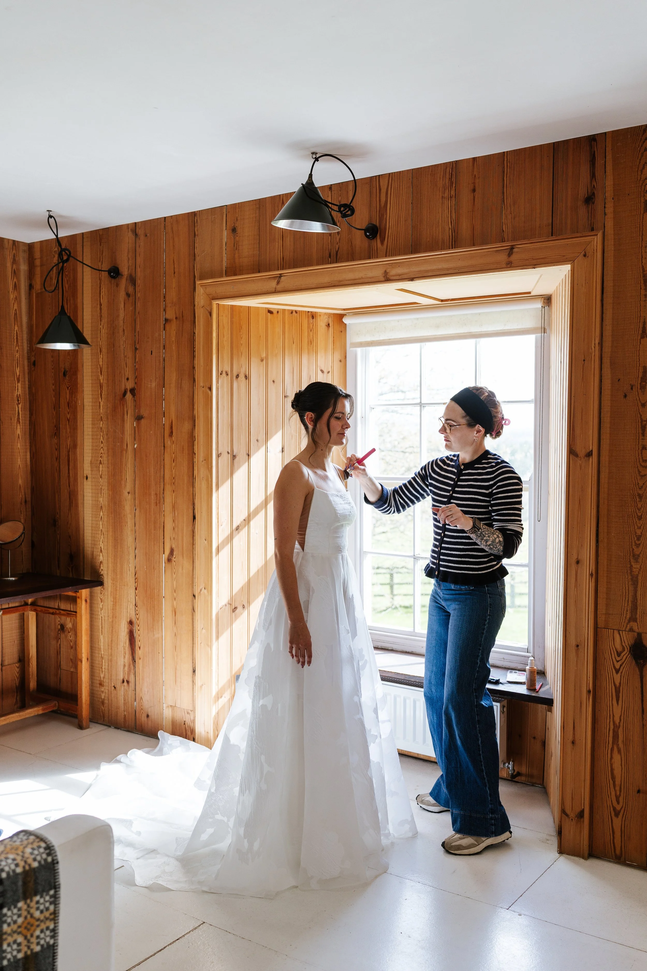 A woman in a wedding dress getting her makeup done by a stylist in a room with wooden walls and a large window.