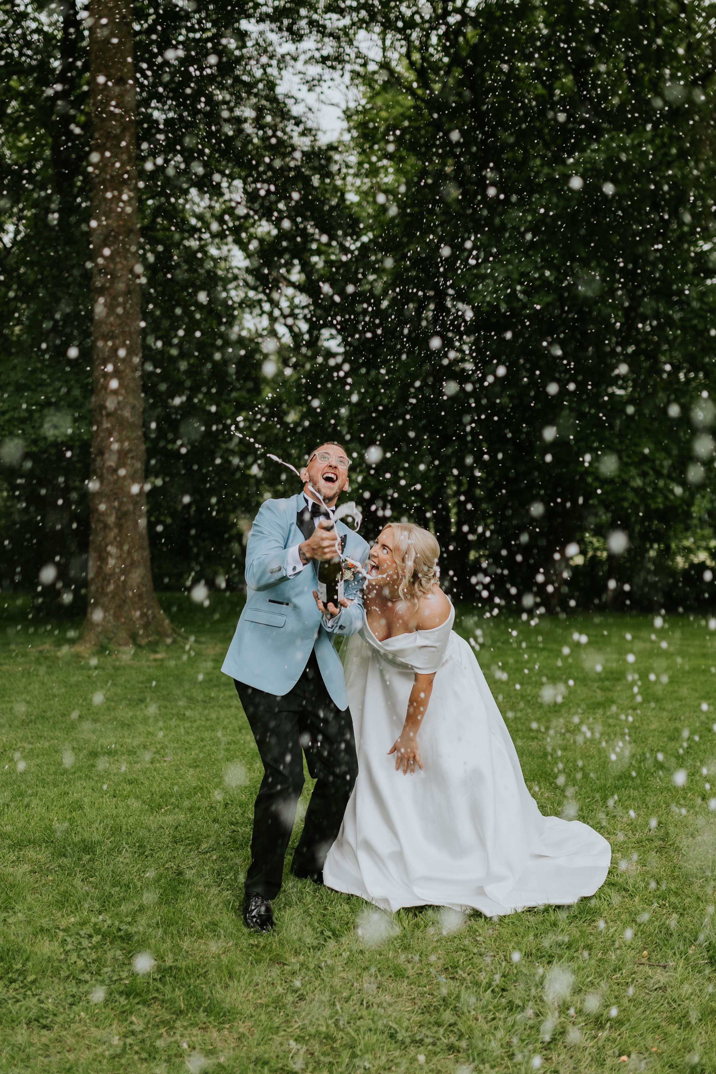 A groom and bride celebrating outdoors on a wedding day, spraying champagne with foam and bubbles in a park with trees.