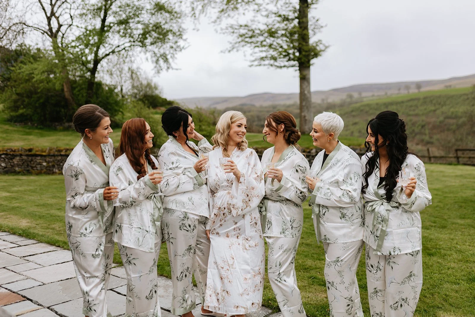 Seven women in floral pajamas, celebrating outdoors with drinks, smiling, with a natural landscape in background.