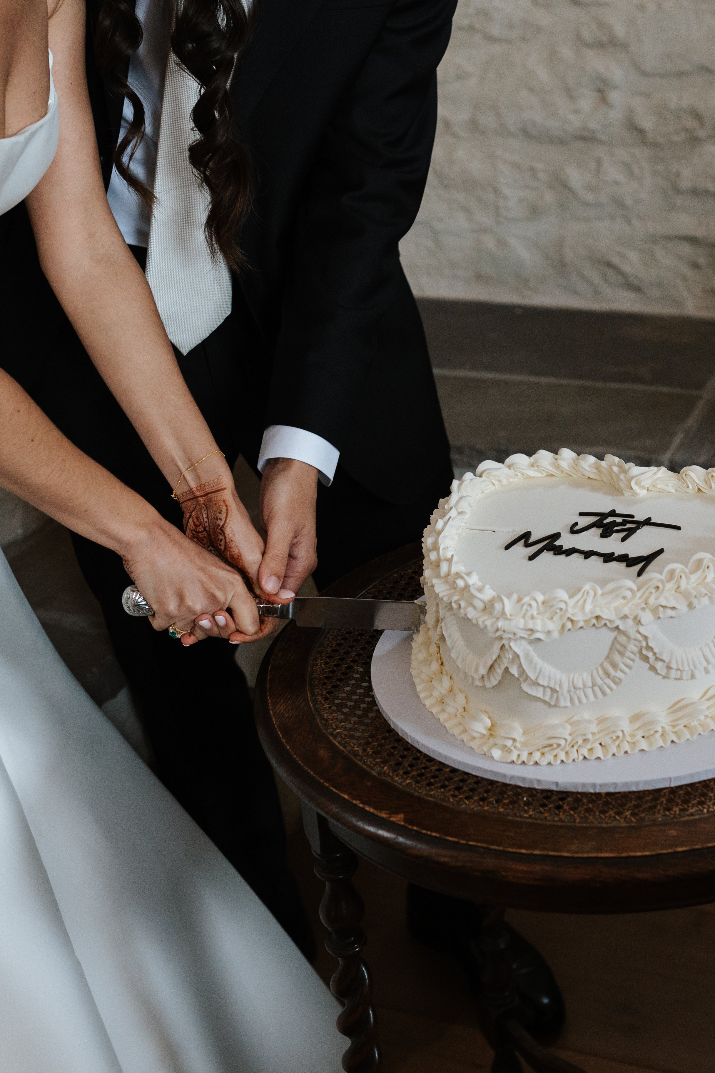 A couple is cutting a wedding cake together. The cake is decorated with white icing and has a message written on it. The groom is wearing a black suit and the bride is in a white dress. They are standing by a dark wooden round table. The bride has he