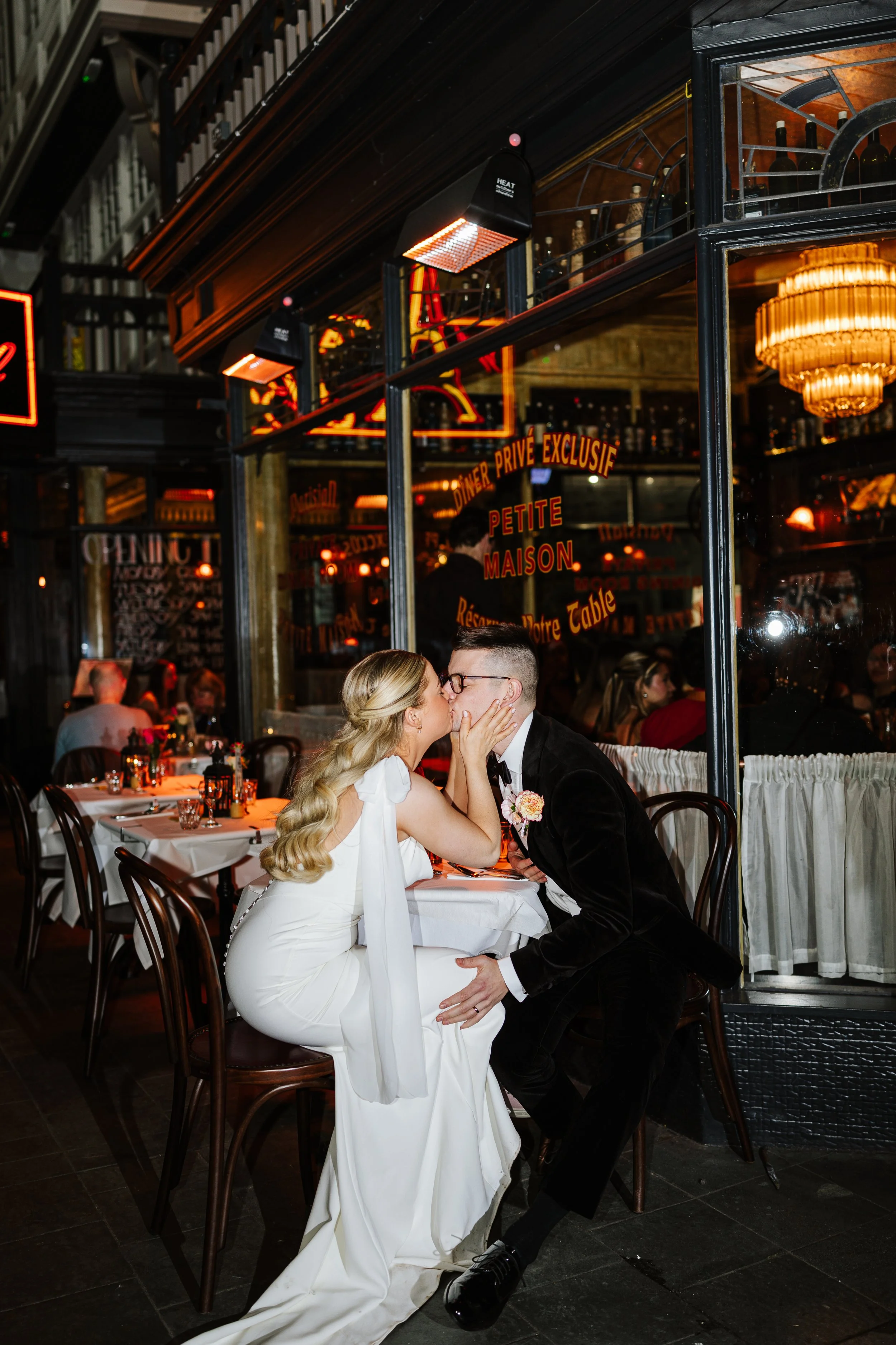 A bride and groom share a kiss in a dimly lit restaurant, with the bride seated and wearing a white dress, and the groom kneeling on one knee.