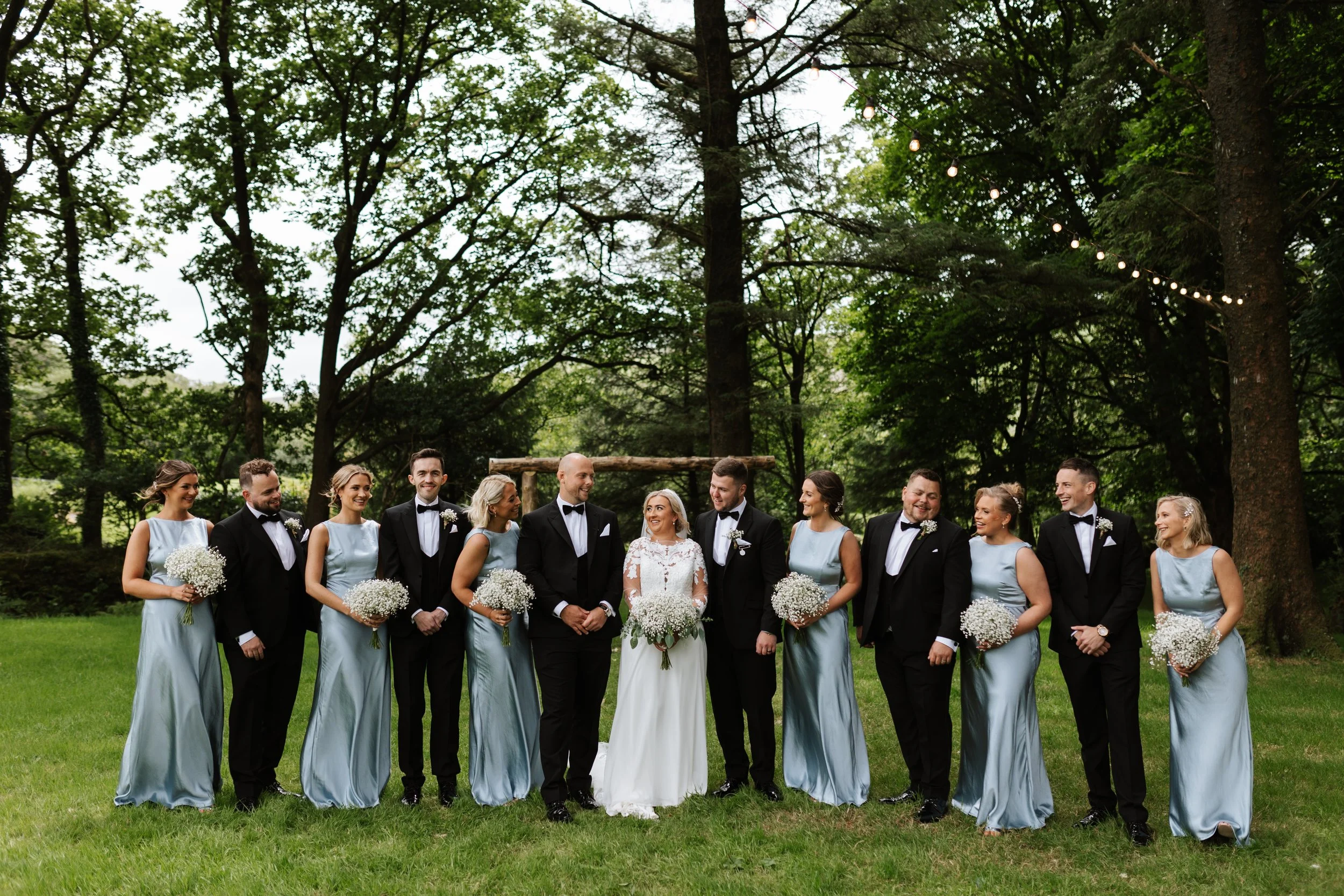 A wedding party standing outdoors on a grassy area surrounded by trees, with string lights hanging overhead. The group includes bridesmaids in matching light blue dresses, groomsmen in black tuxedos, and a bride in a white wedding gown holding a bouq