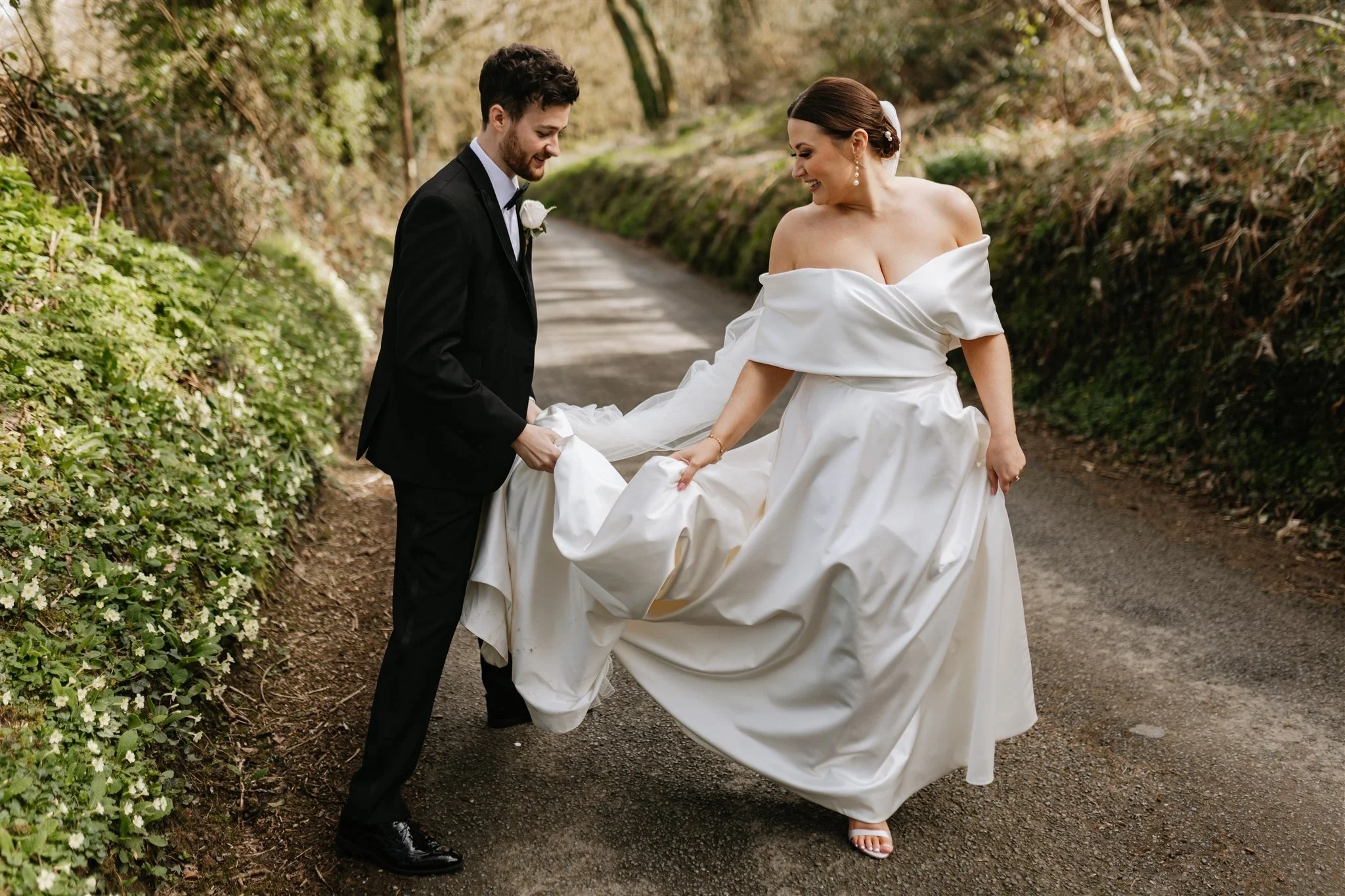 A bride and groom, dressed in wedding attire, sharing a joyful moment outdoors on a wooded path. The groom is in a black tuxedo and the bride in a white off-shoulder wedding gown, lifting her dress as they smile at each other.