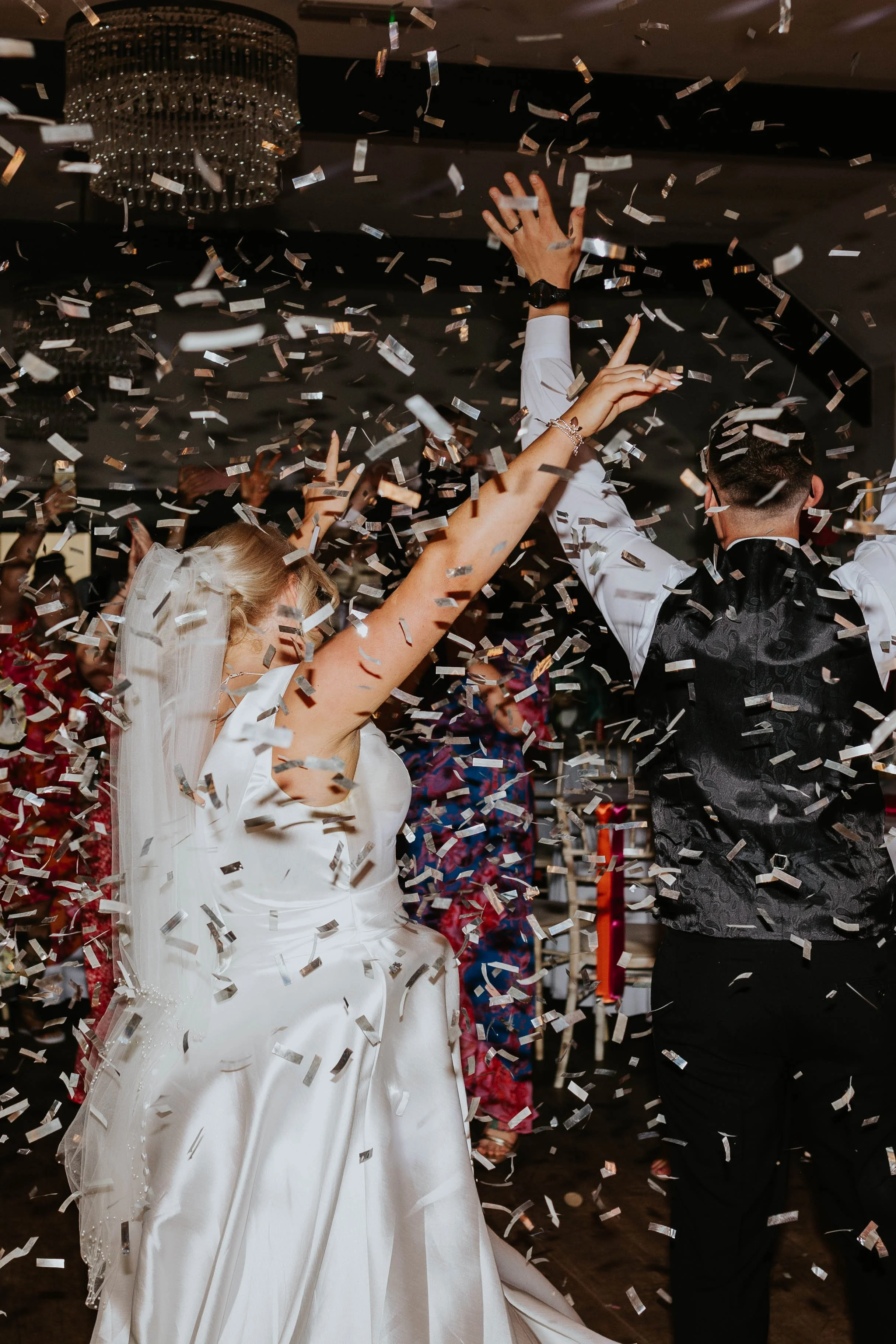 A bride in a white wedding dress and a groom in a black vest and white shirt are dancing under a shower of silver confetti at a celebration, with guests in the background.