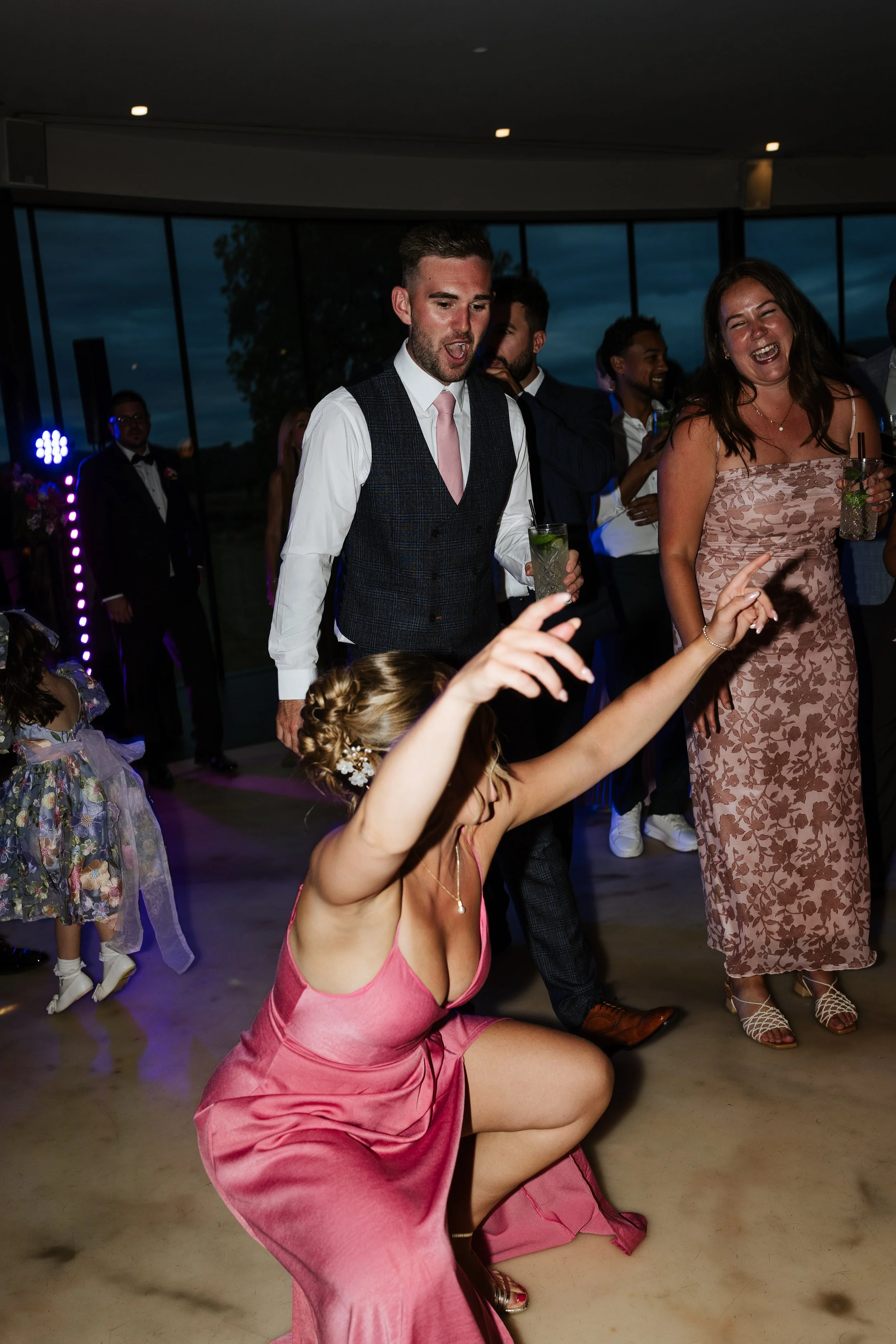 People dancing and celebrating at a wedding reception indoors with large windows showing a sunset sky. A woman in a pink dress is kneeling and dancing in the foreground, while others are standing and smiling in the background.