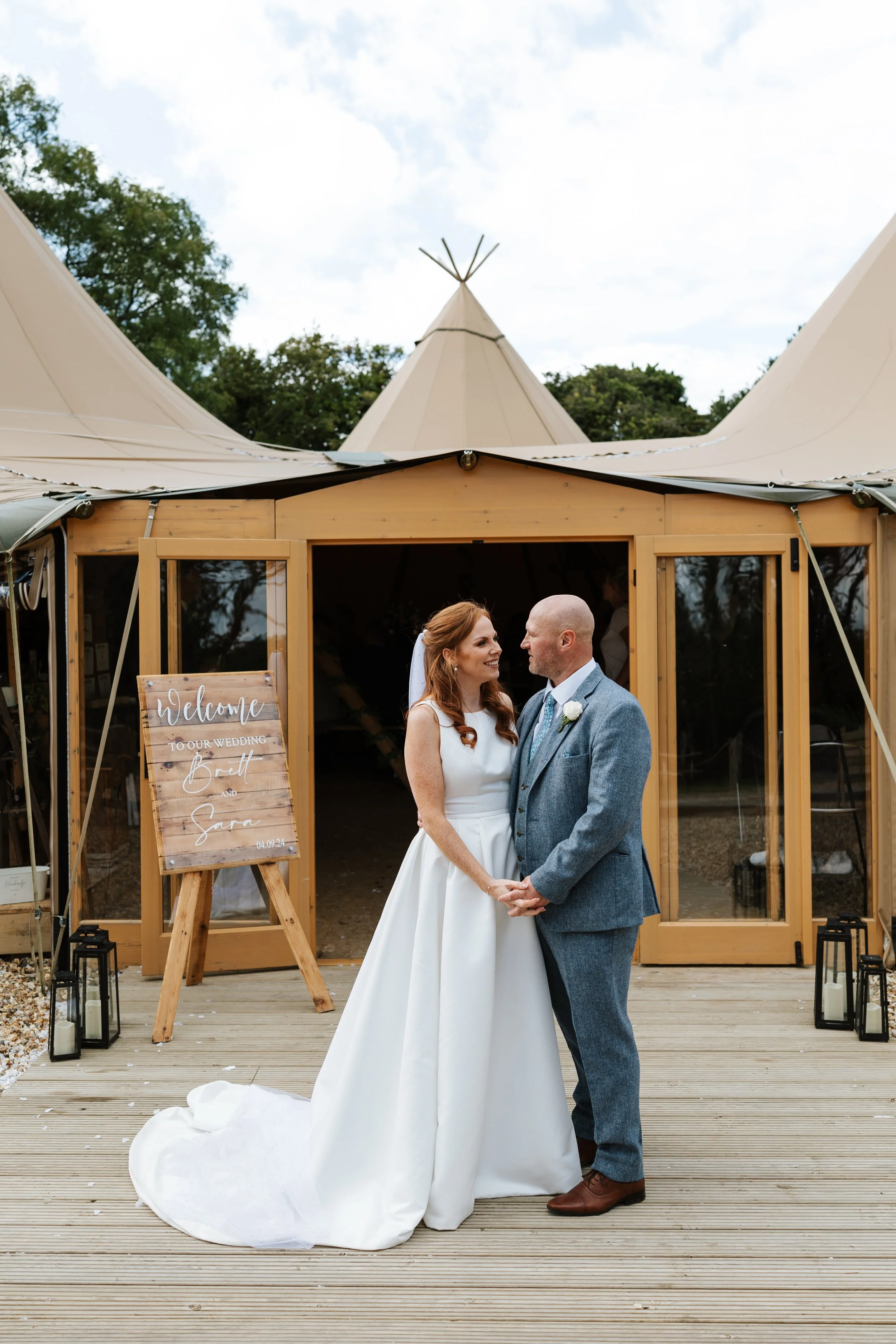 Bride and groom standing hand-in-hand outside a wedding venue with a wooden gateway, a sign welcoming guests to their wedding, and a tipi tent in the background.