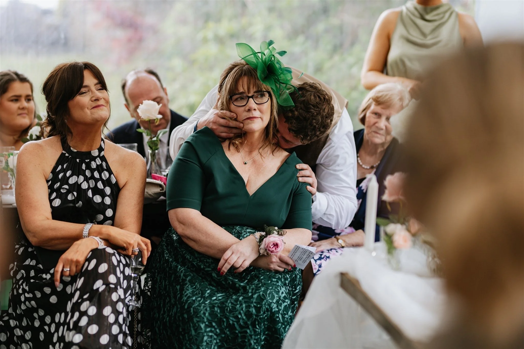 People attending a wedding ceremony, with one woman in a green dress and a green fascinator, sitting with her arms crossed, while a man behind her embraces her with a kiss on the cheek. Other guests are seated nearby, wearing formal attire.