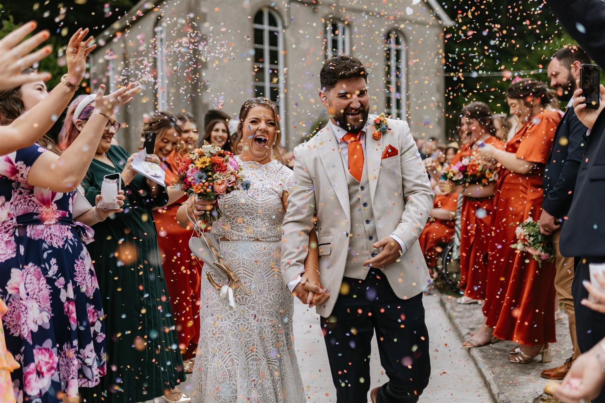 A newly married couple holding hands and laughing as they walk through a crowd of guests throwing colorful confetti during an outdoor wedding celebration.