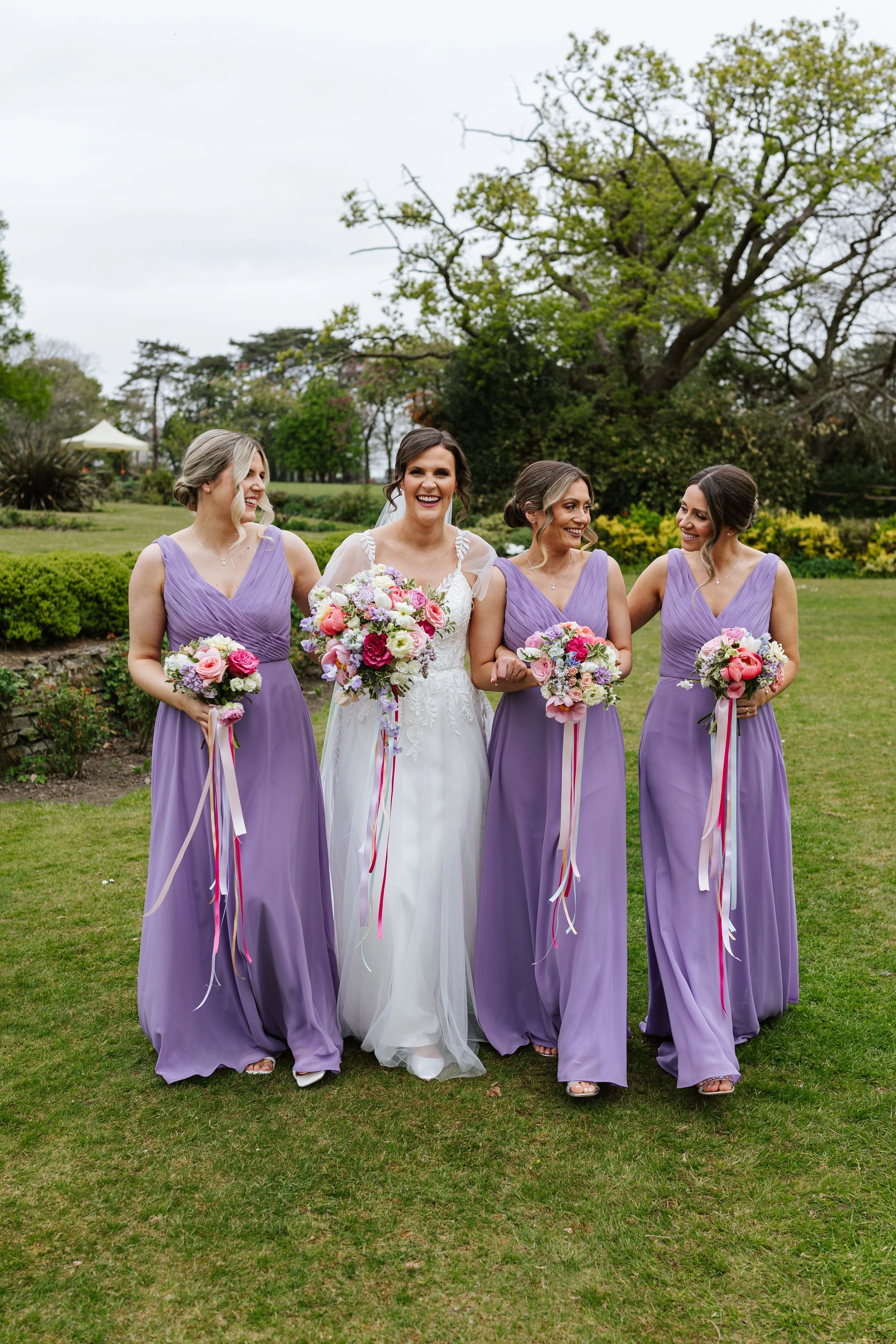 Bride in white wedding dress standing between four bridesmaids in lavender dresses holding bouquets of pink, purple, and white flowers, in a garden with trees and greenery.