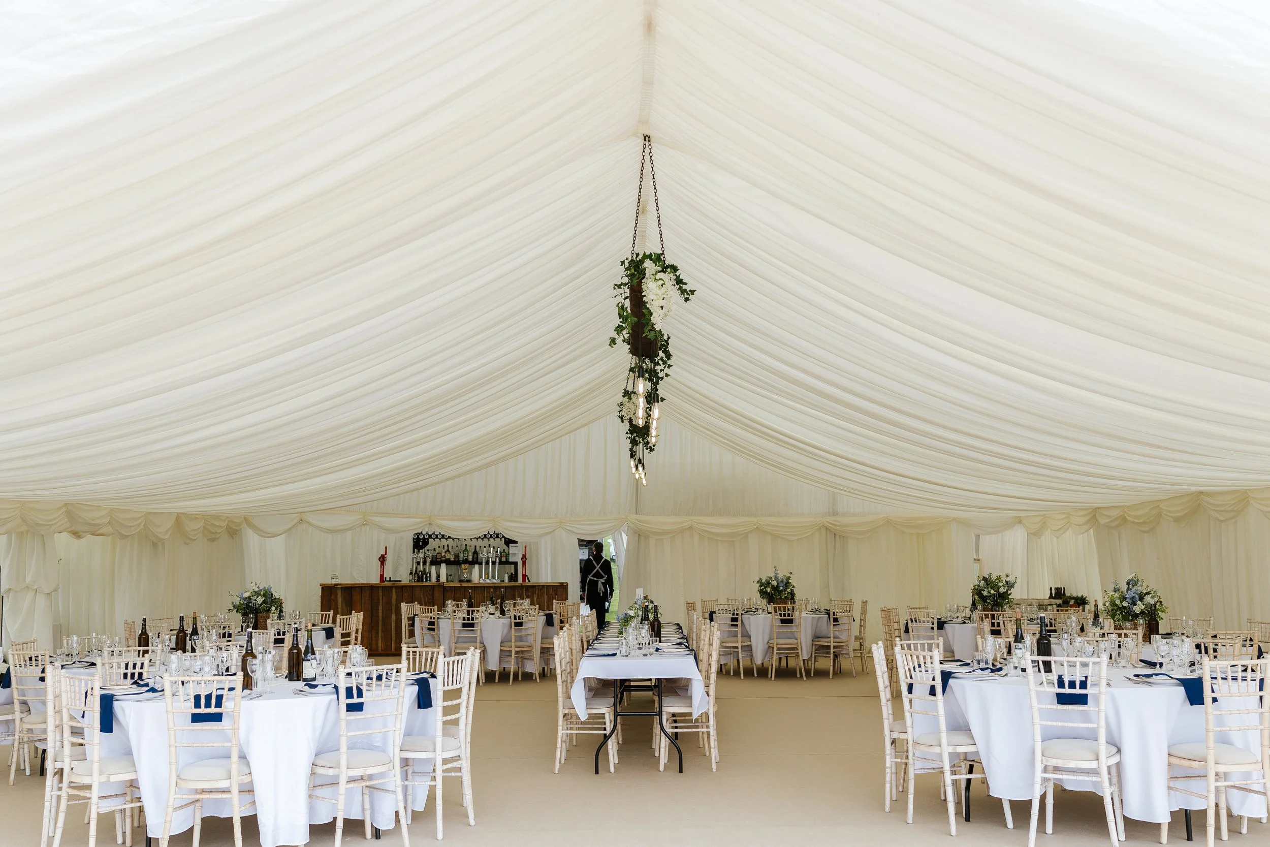 Interior of a large, elegant event tent with white draped ceiling and hanging chandelier decorated with greenery and white flowers. Round tables with white tablecloths, set with glassware, bottles, and blue napkins, and surrounded by white chairs. In