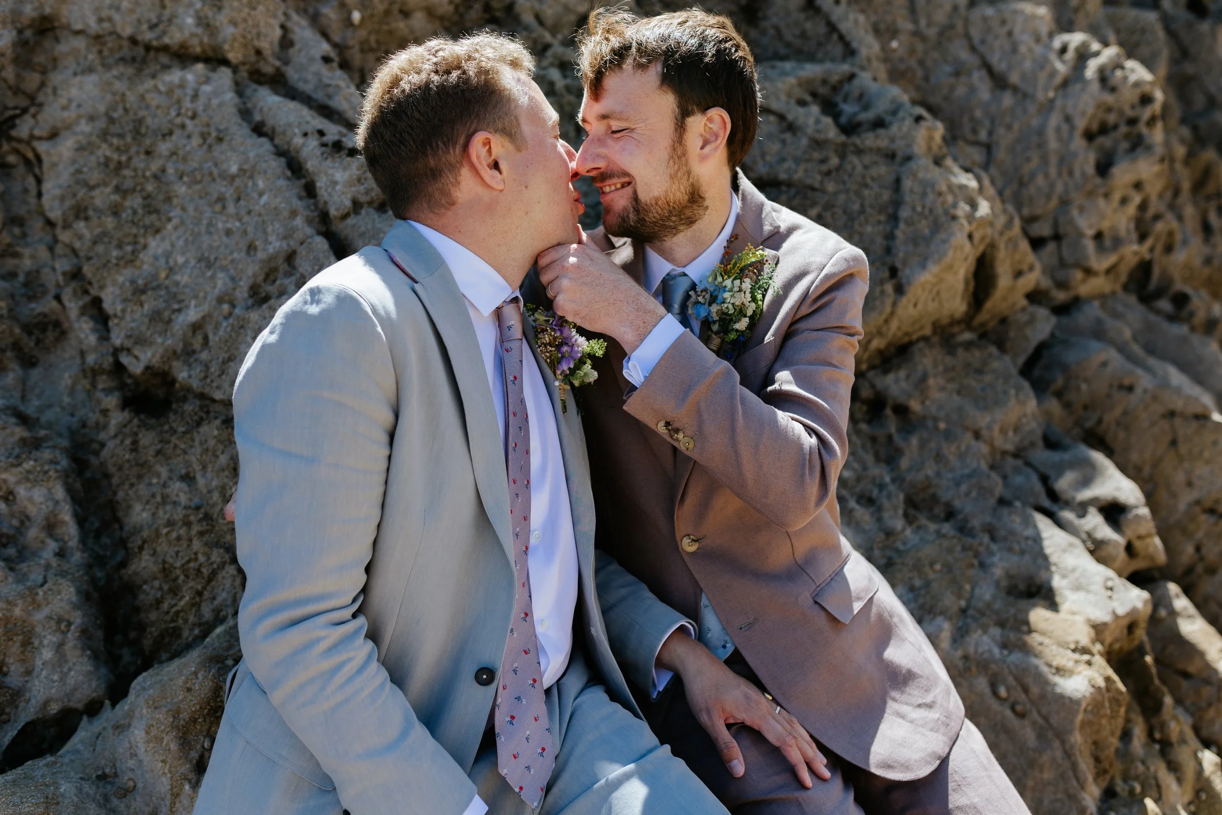 Two men in suits sharing an intimate moment on a rocky outdoor setting, smiling and touching faces.