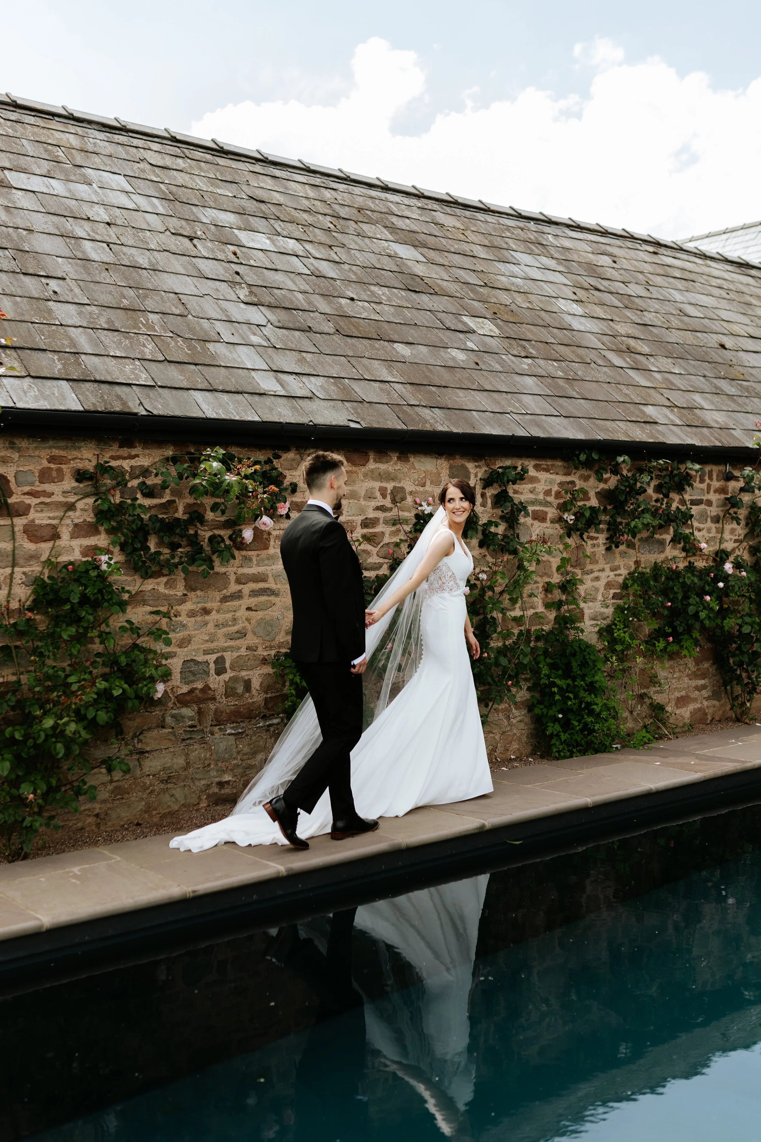 A bride and groom standing on a stone pathway beside a swimming pool with a brick wall and greenery in the background. The bride is wearing a white wedding dress with a long train and veil, smiling at the groom. The groom is dressed in a black suit, 
