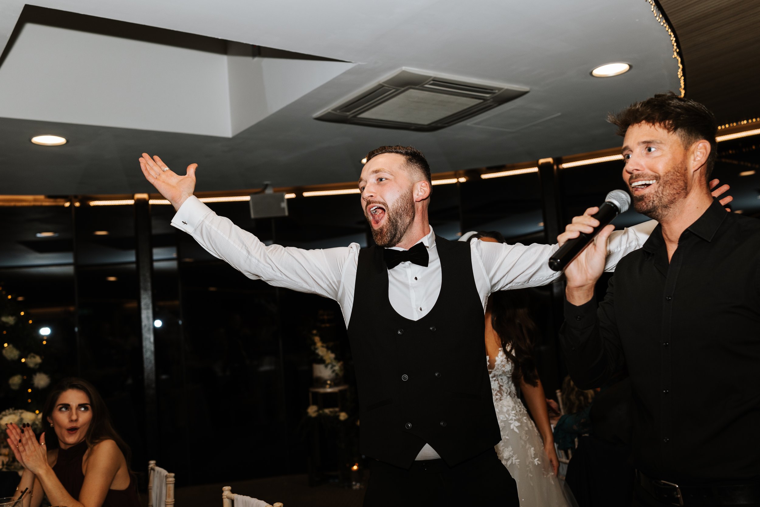 Two men, dressed formally, are standing and singing into a microphone at a celebration or wedding reception, with one man raising his arm expressive and joyful. A woman is seated nearby, clapping and looking surprised or excited.