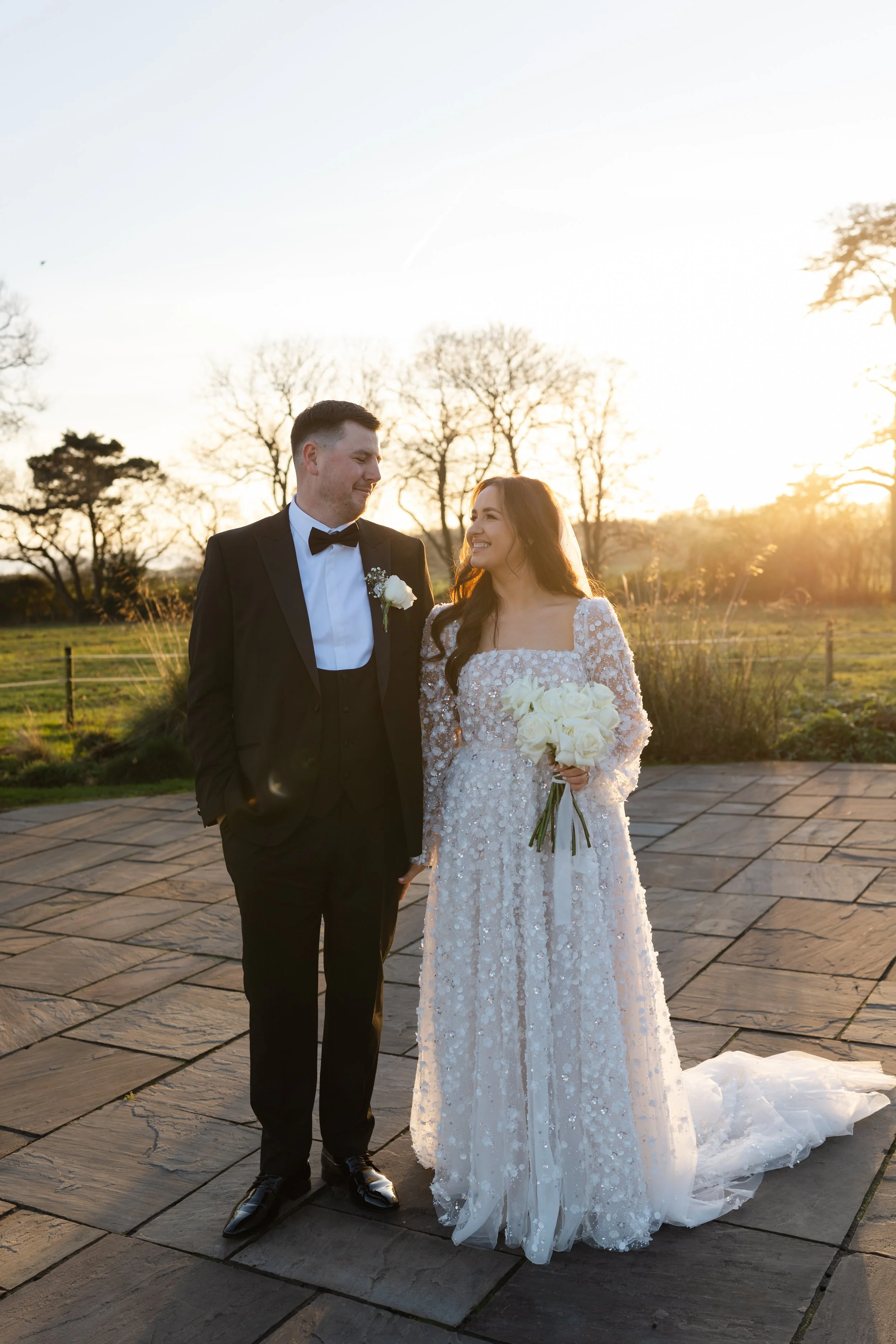 A bride and groom stand outdoors during sunset, smiling at each other. The bride wears a white, lace wedding gown holding a bouquet of white flowers, and the groom wears a black tuxedo with a bow tie and a boutonniere. Bare trees and a grassy field a